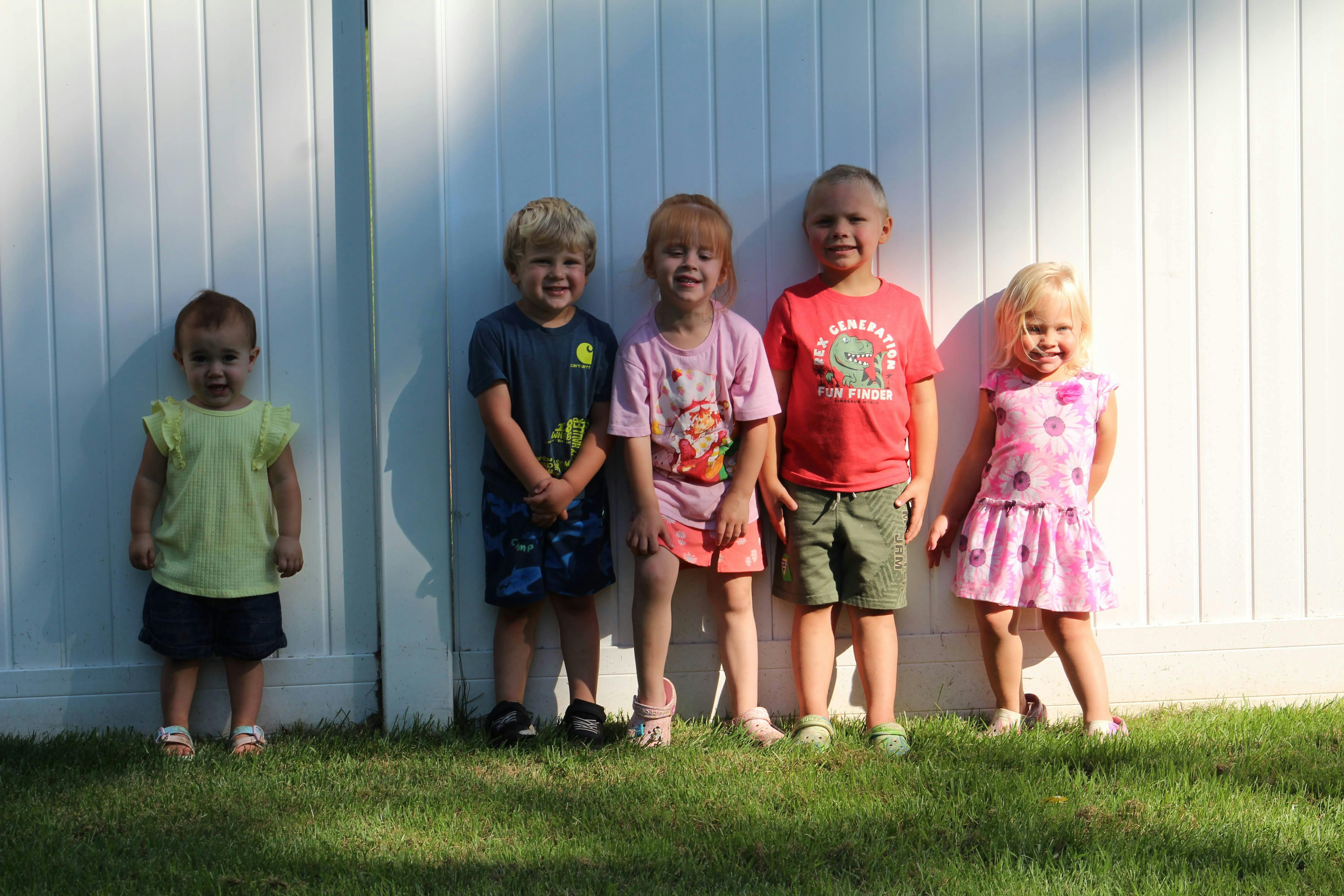 Five children standing in a row against a white fence