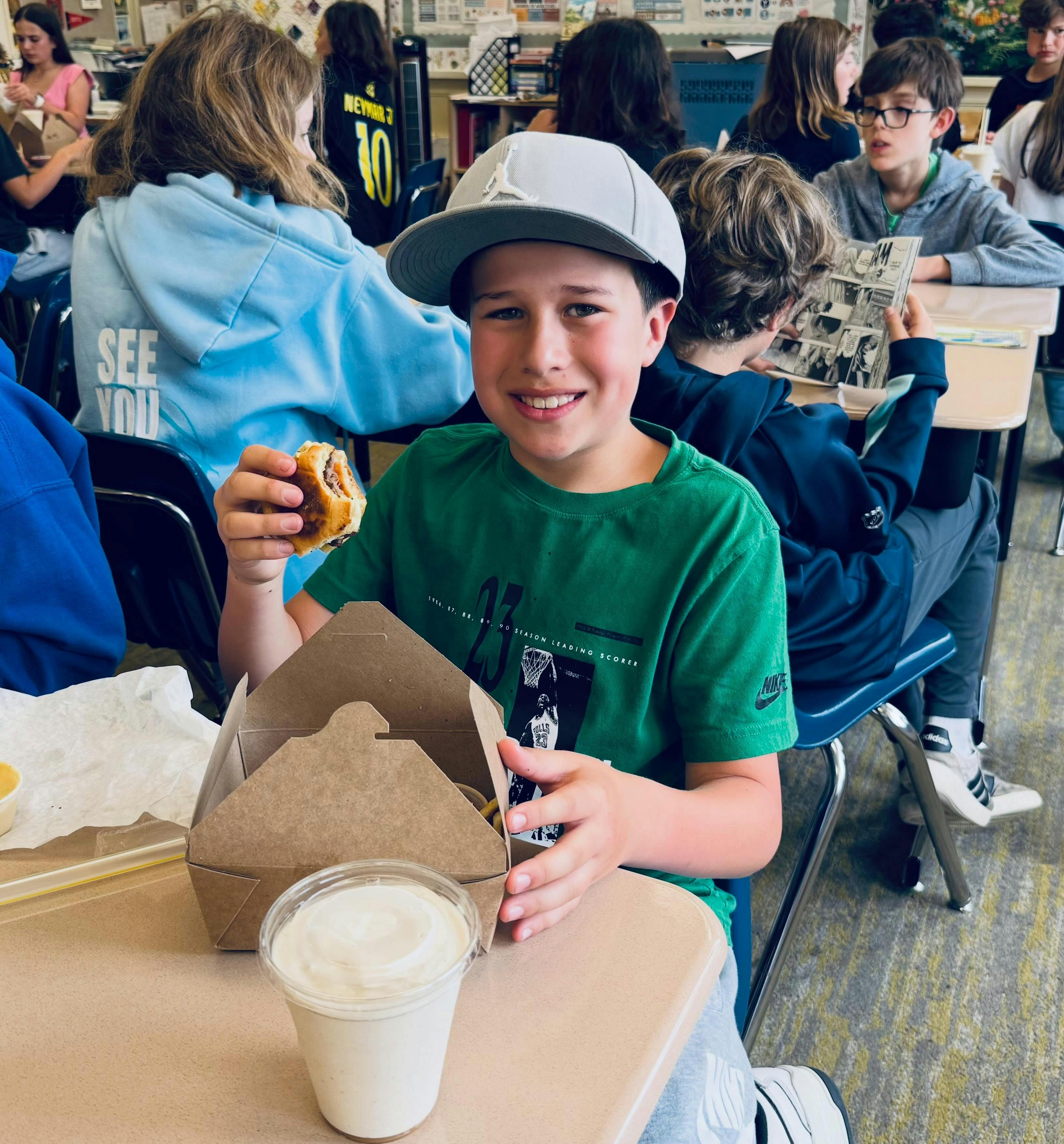 Child smiling while holding a half-eaten sandwich and a drink in a classroom