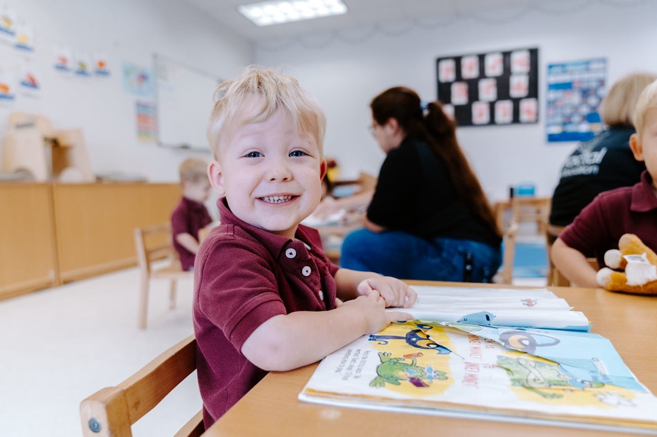 Child smiling at a desk with an open book in a classroom