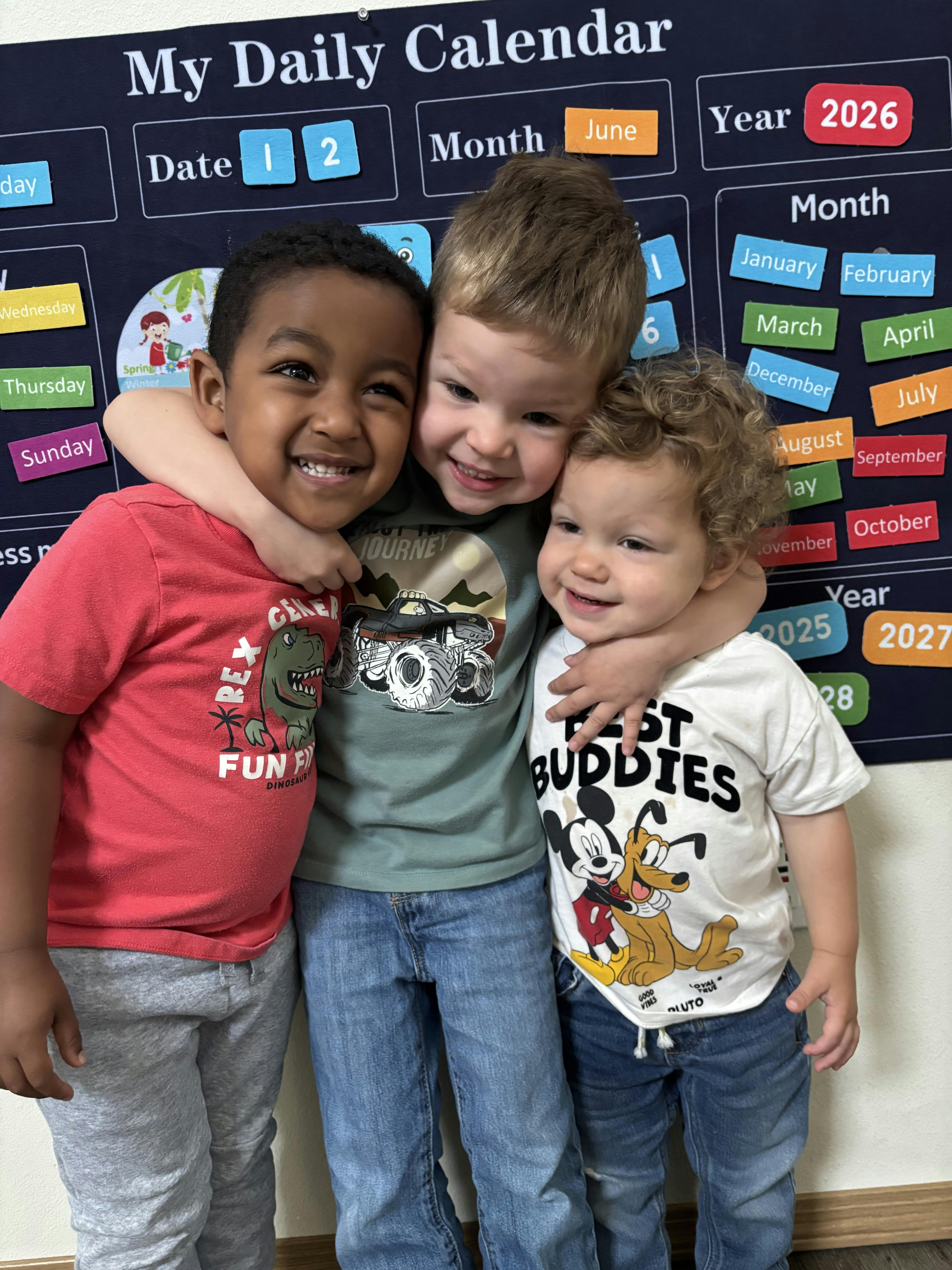 Three smiling young children hugging in front of a daily calendar board