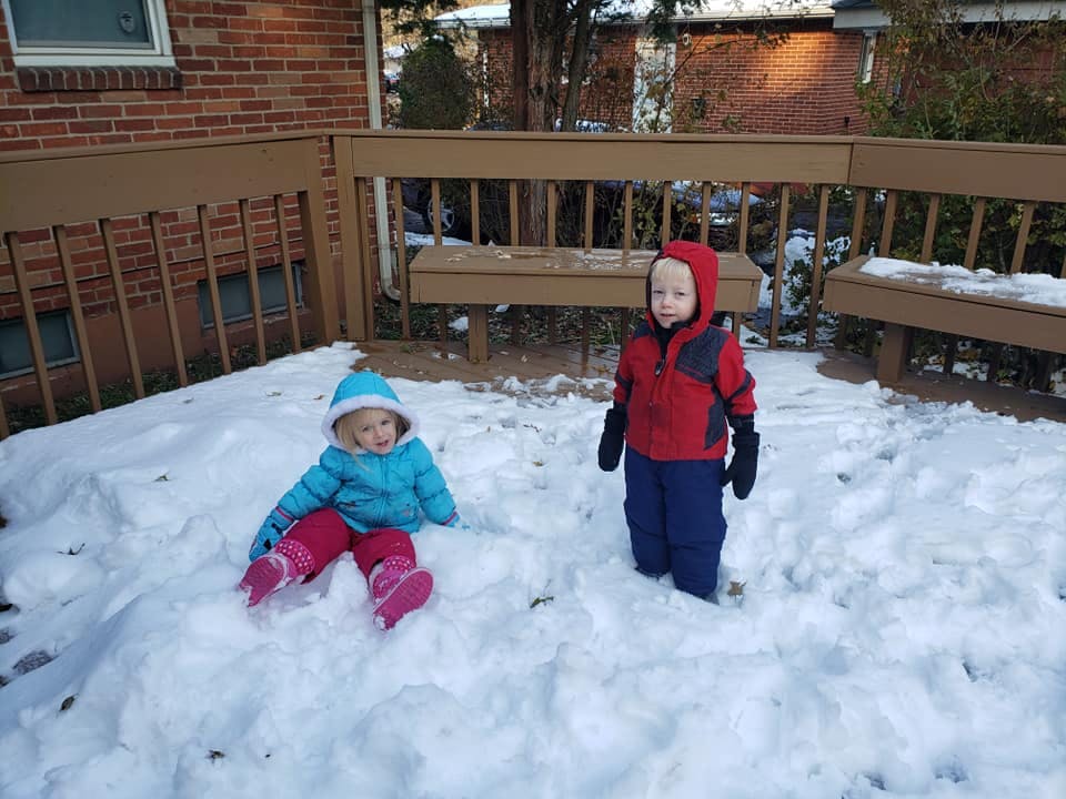 Two children in winter clothes playing in snow on a snowy deck