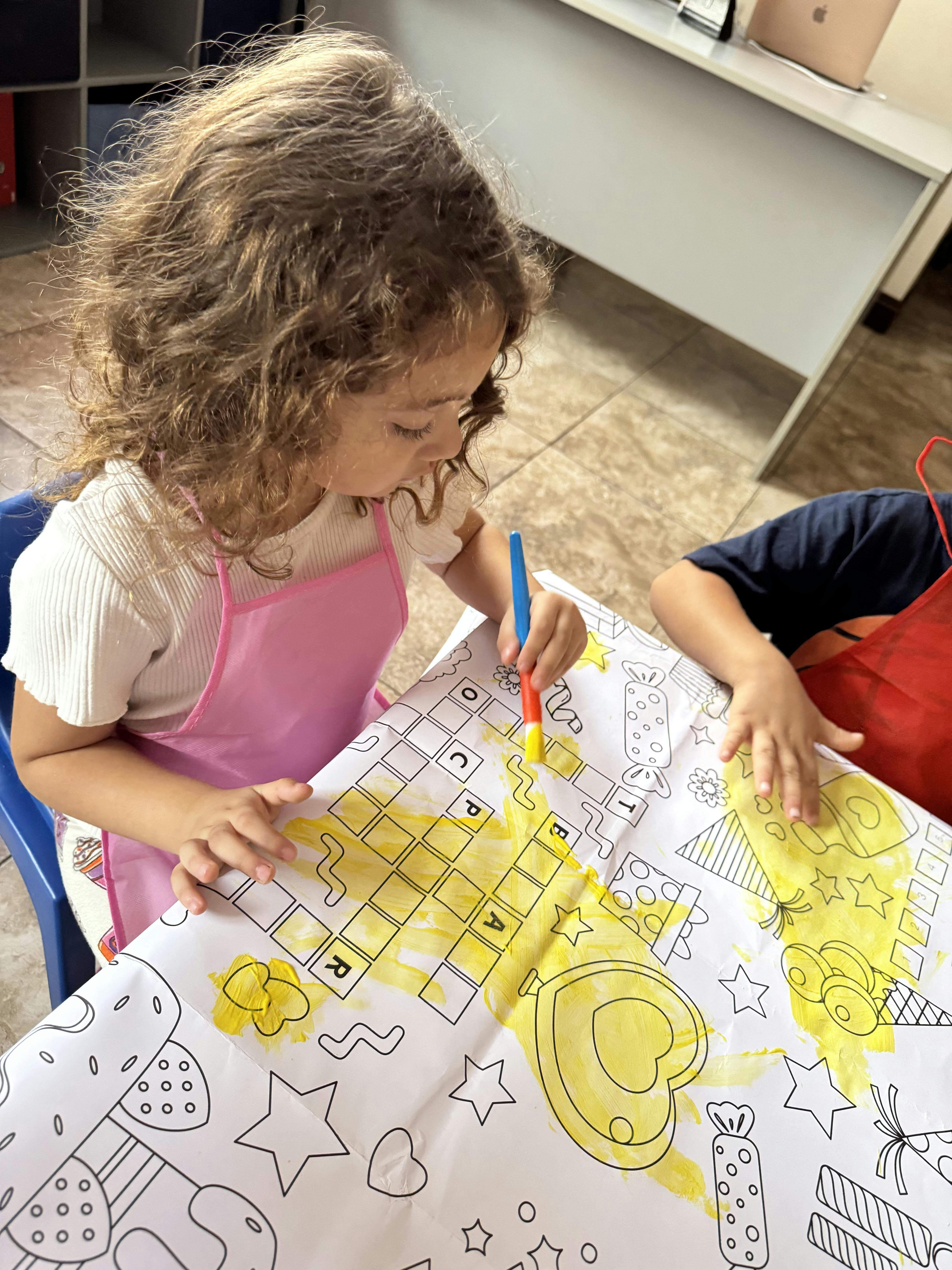 Child in pink apron painting on a large paper with yellow paint