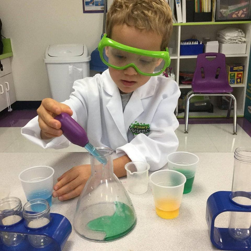 Child in lab coat and safety goggles using a dropper with beakers