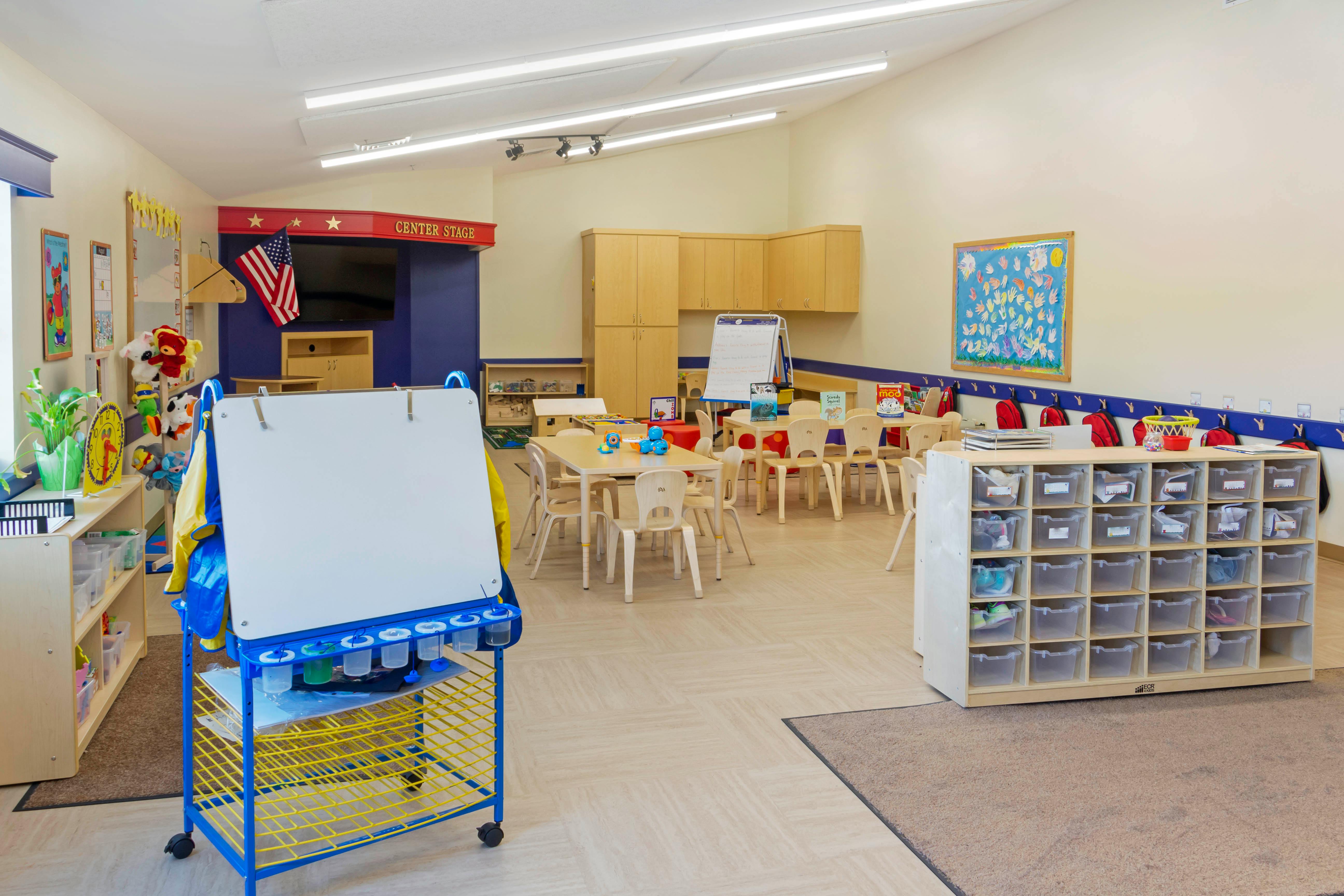 An empty, bright children's classroom with small tables and chairs, shelves, and cubbies