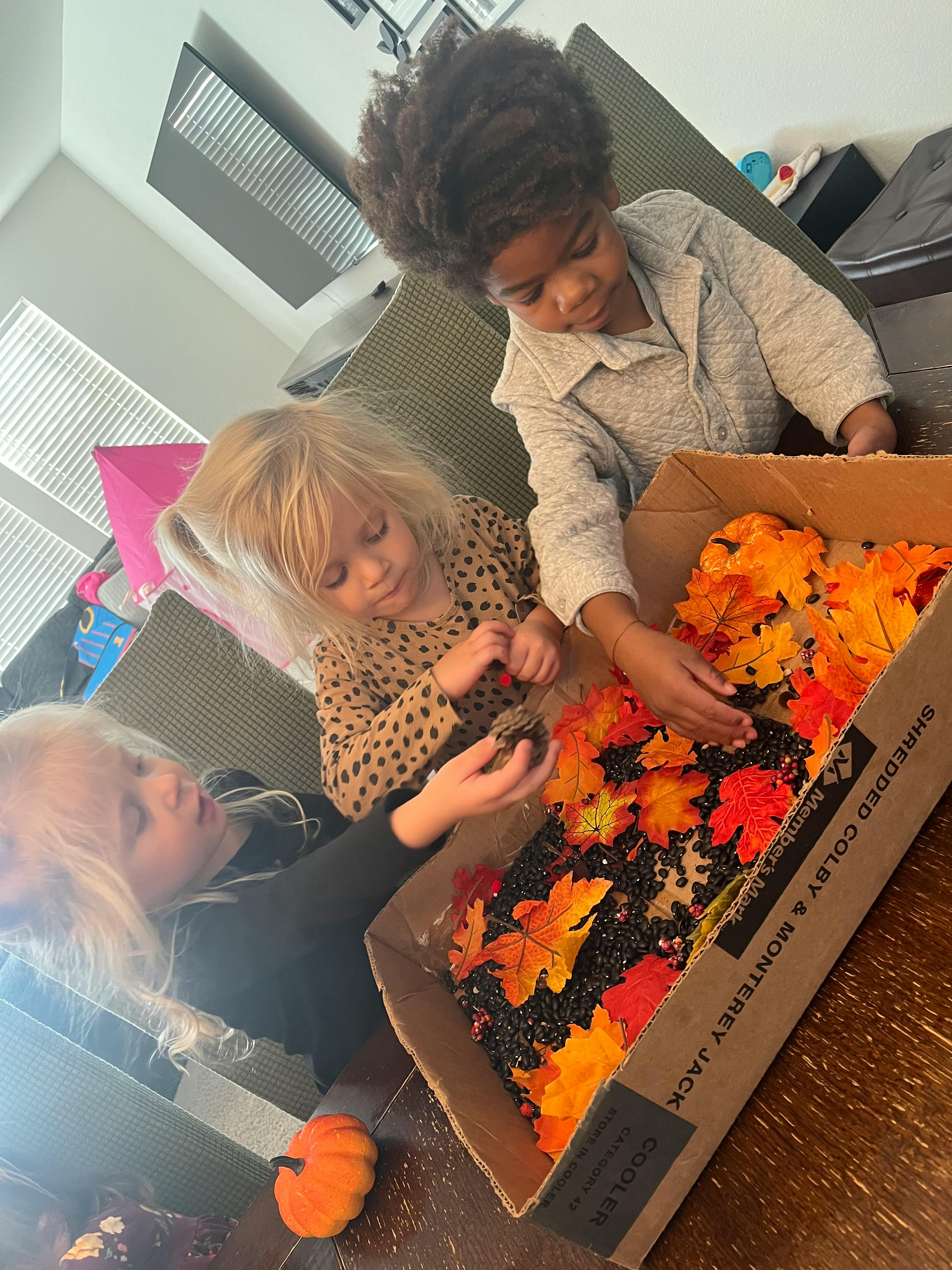 Three children playing with fall leaves and small black objects in a box