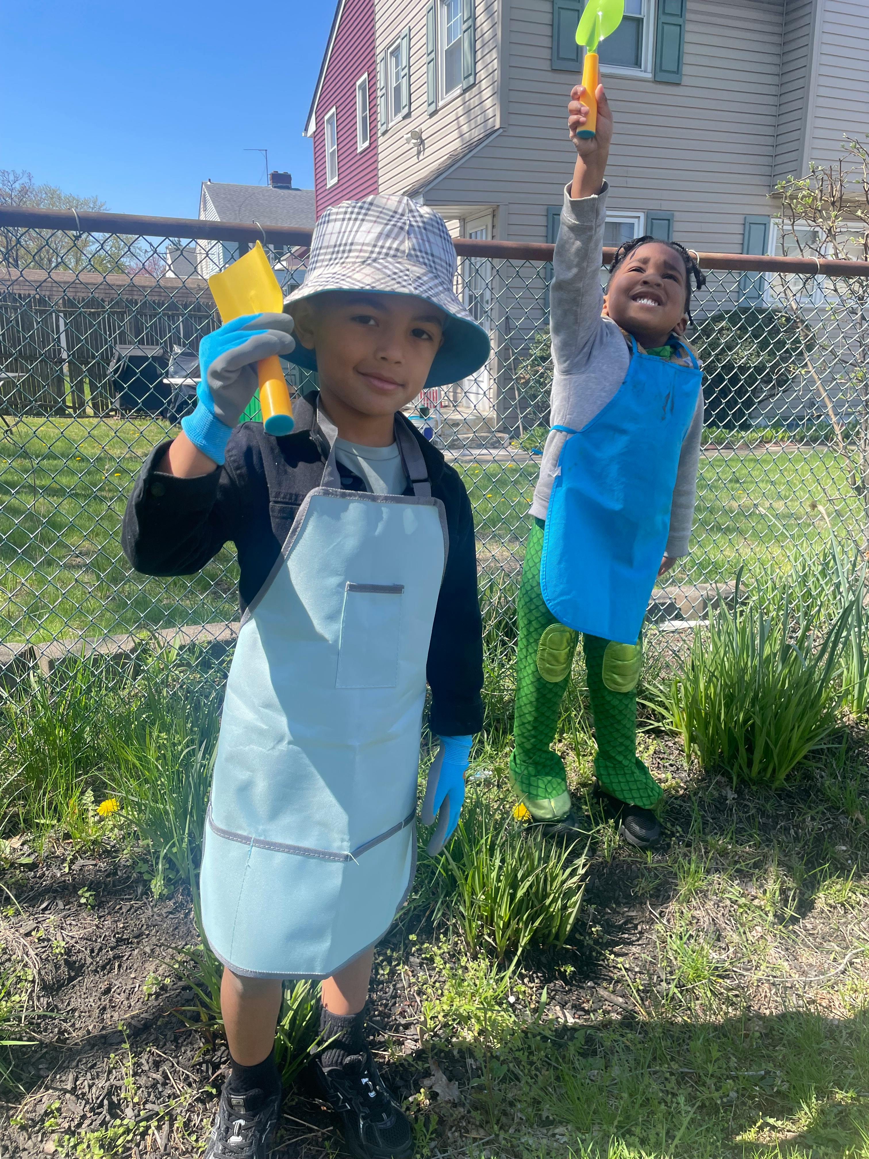 Two children wearing aprons and gloves, holding gardening tools