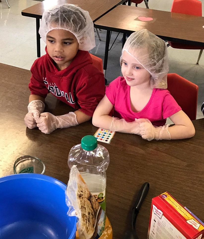 Two children wearing hairnets and gloves at a table with cooking ingredients