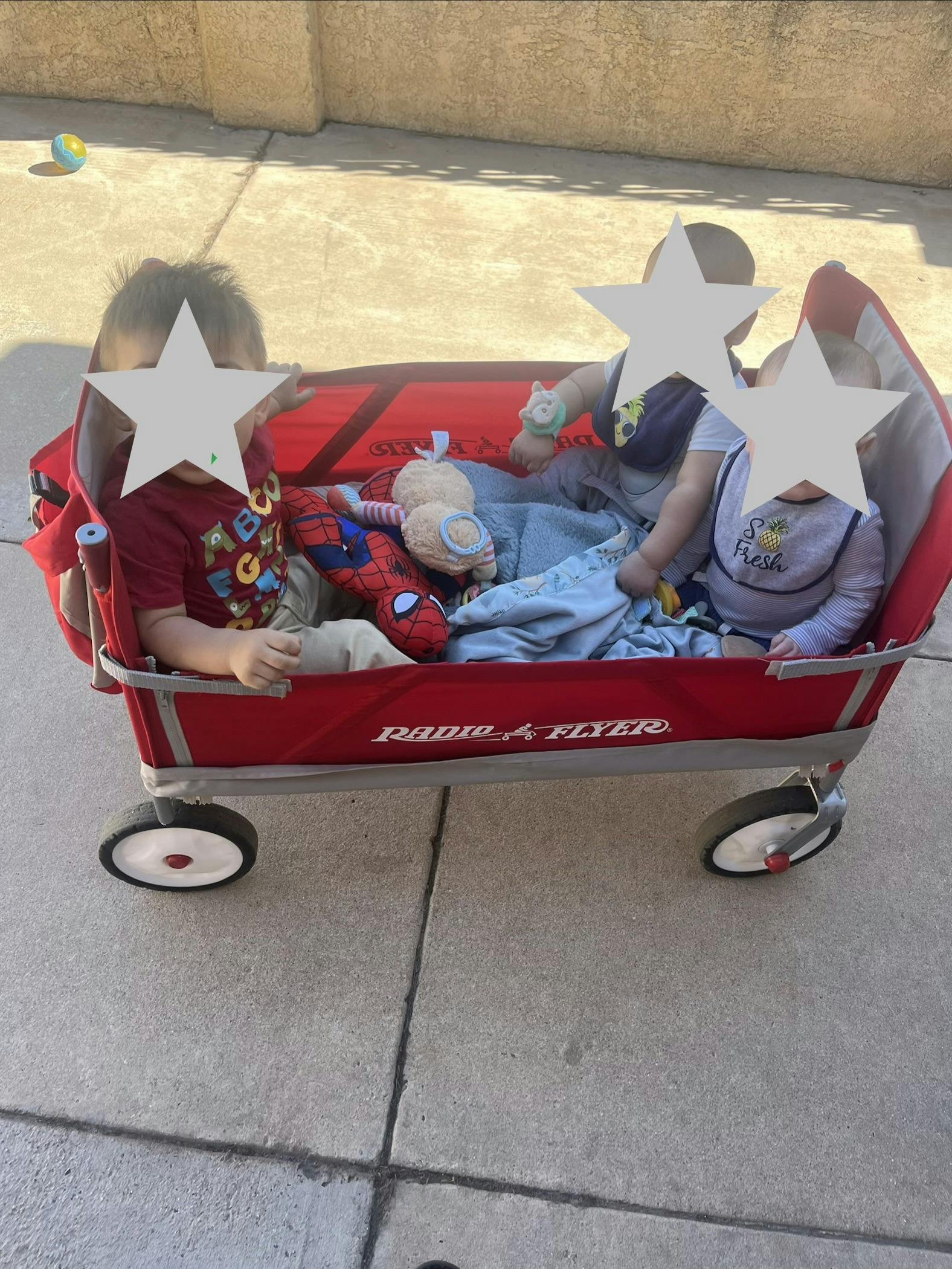 Three children sitting in a red wagon with stuffed animals and blankets