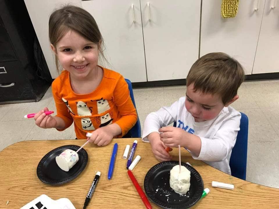 Two children decorating white objects on sticks at a table