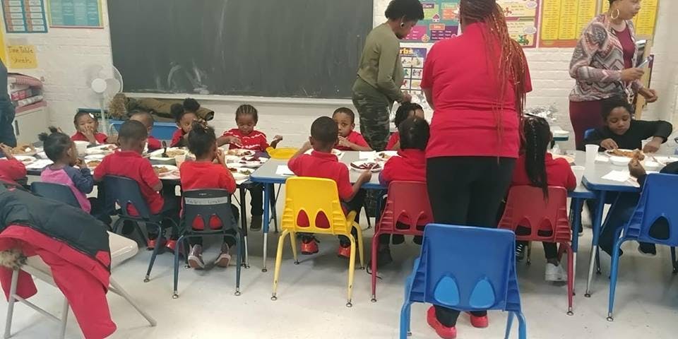 Adults assisting children seated at tables in a classroom