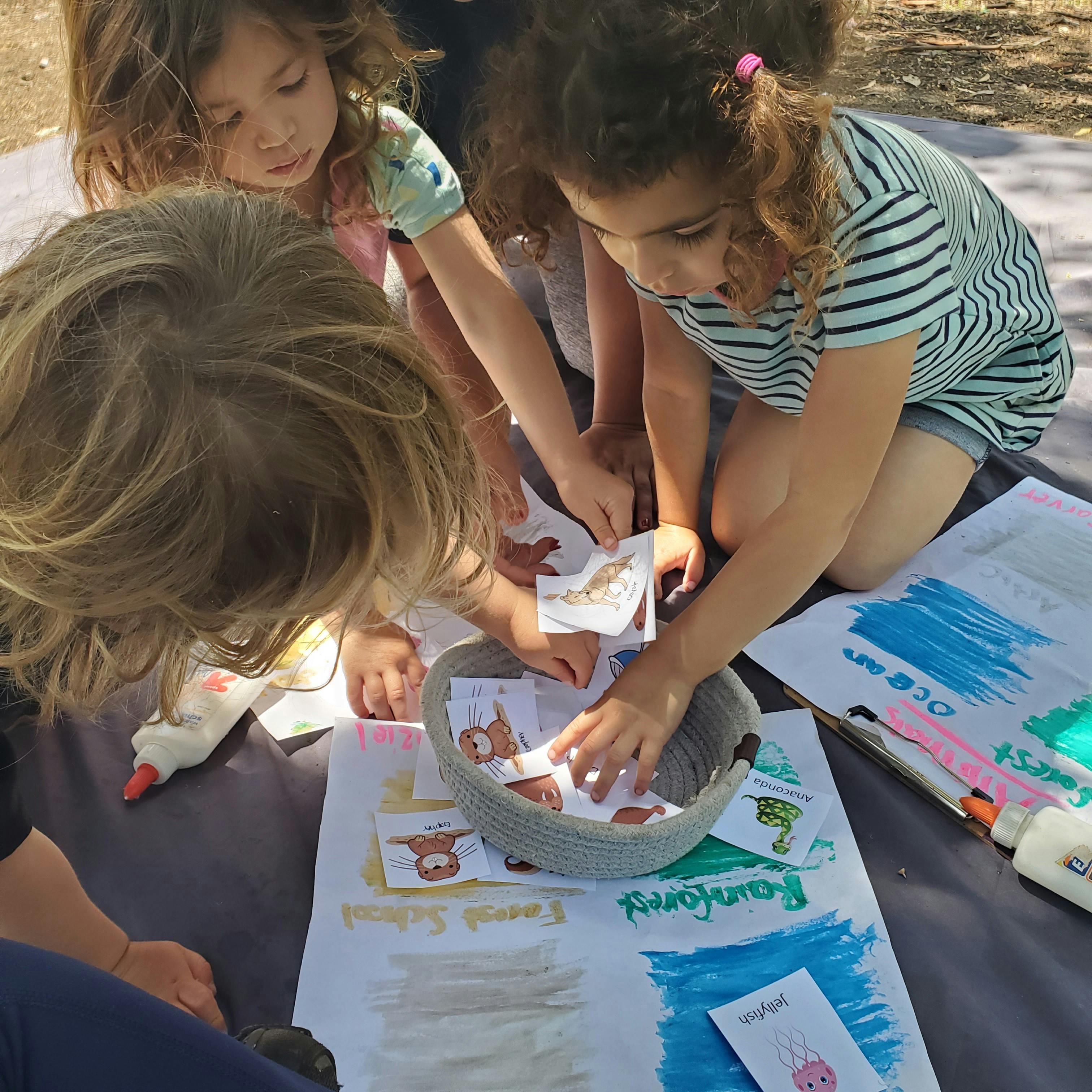 Three children sorting small cards into a bowl on a blanket