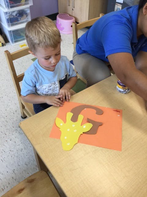 Child at a table with a craft project of a giraffe head and the letter G