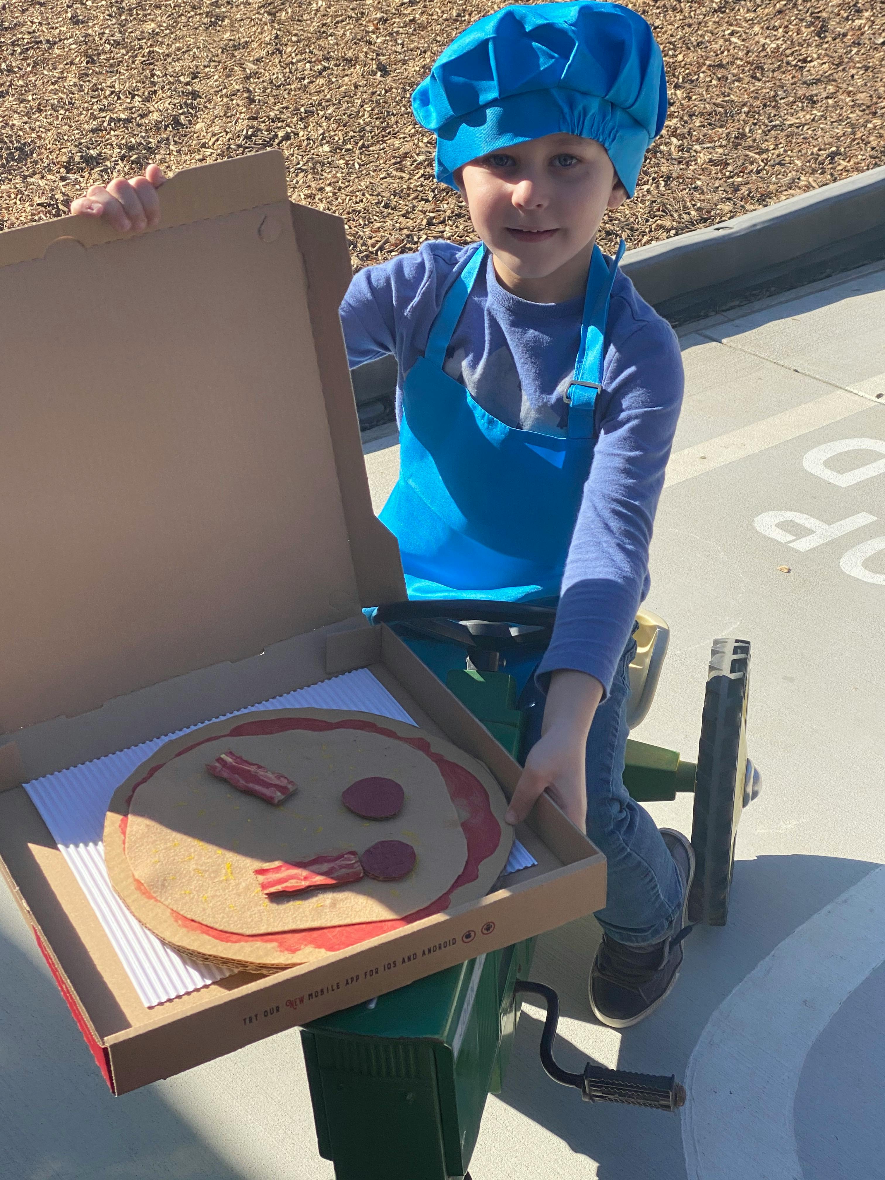 Child in chef hat and apron holding a pizza box with a toy pizza inside