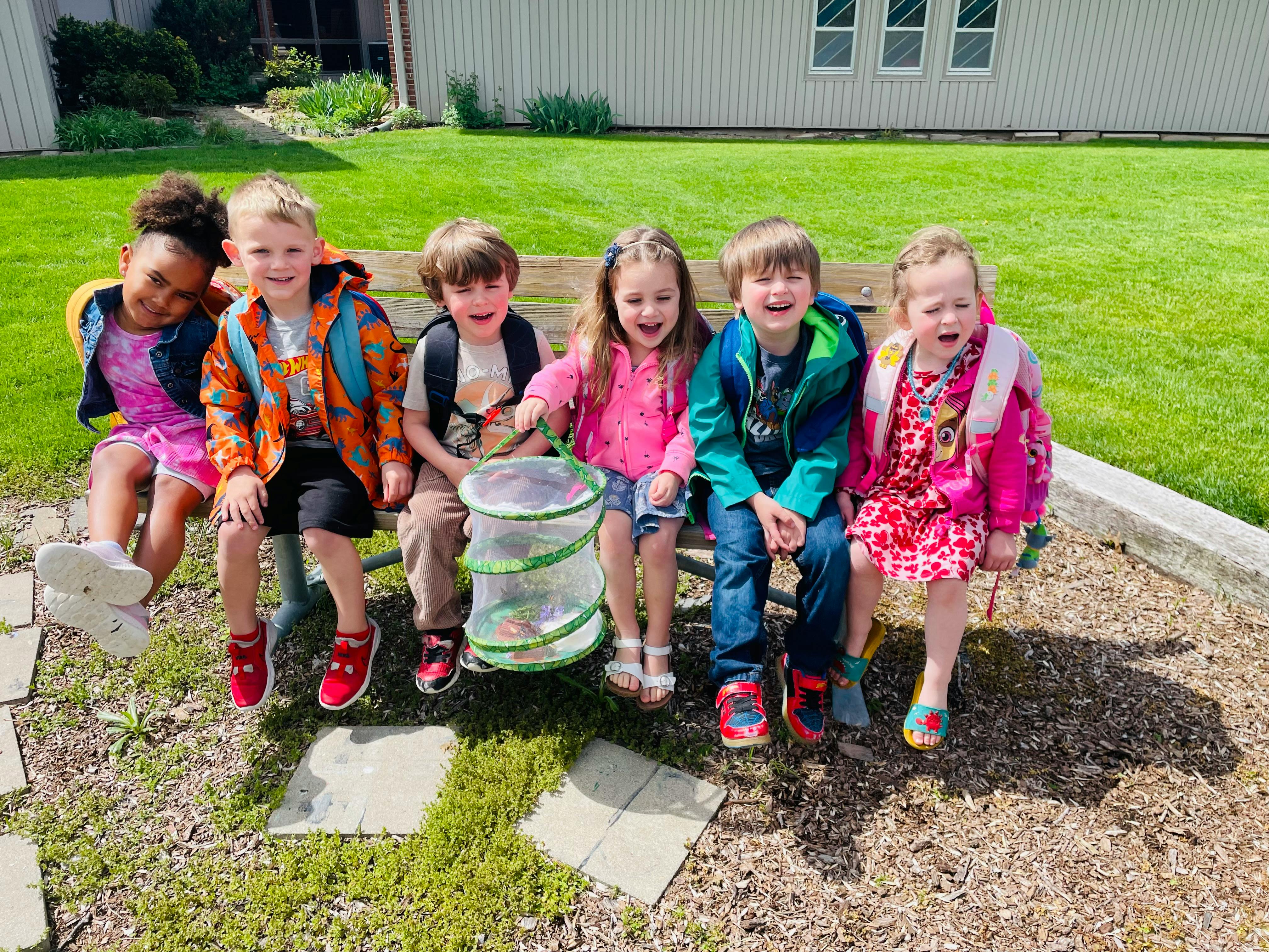 Five children sitting on a bench outdoors with a bug habitat