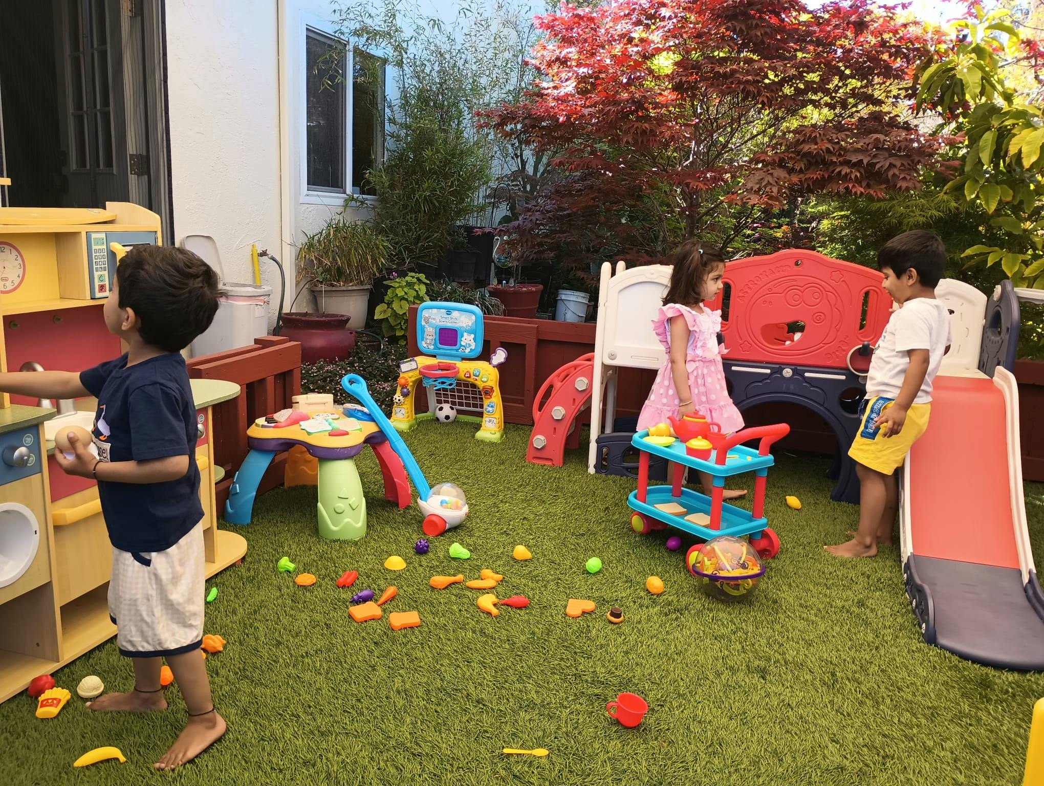 Three children playing outdoors with toy kitchen and slide on artificial turf
