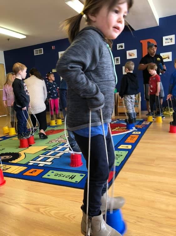 Child walking on bucket stilts in a classroom with other children