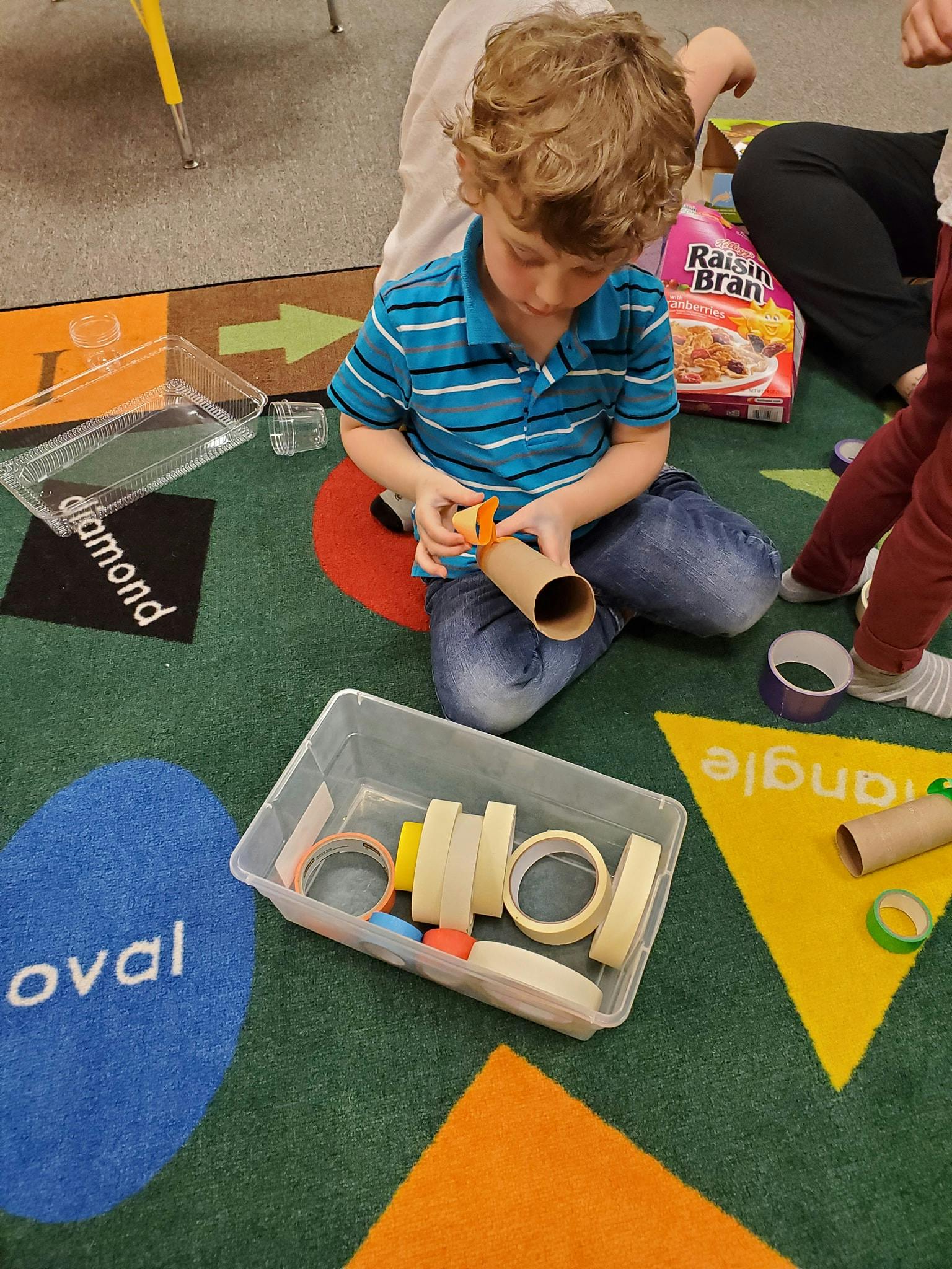 Child kneeling on floor, holding a cardboard tube and tape