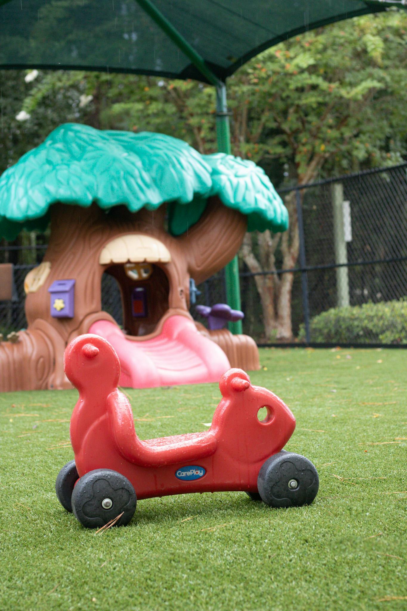 Red ride-on toy on artificial grass with a playhouse in the background