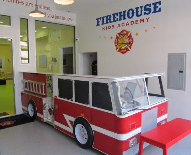 Red and white fire truck play structure indoors with "FIREHOUSE KIDS ACADEMY" on wall