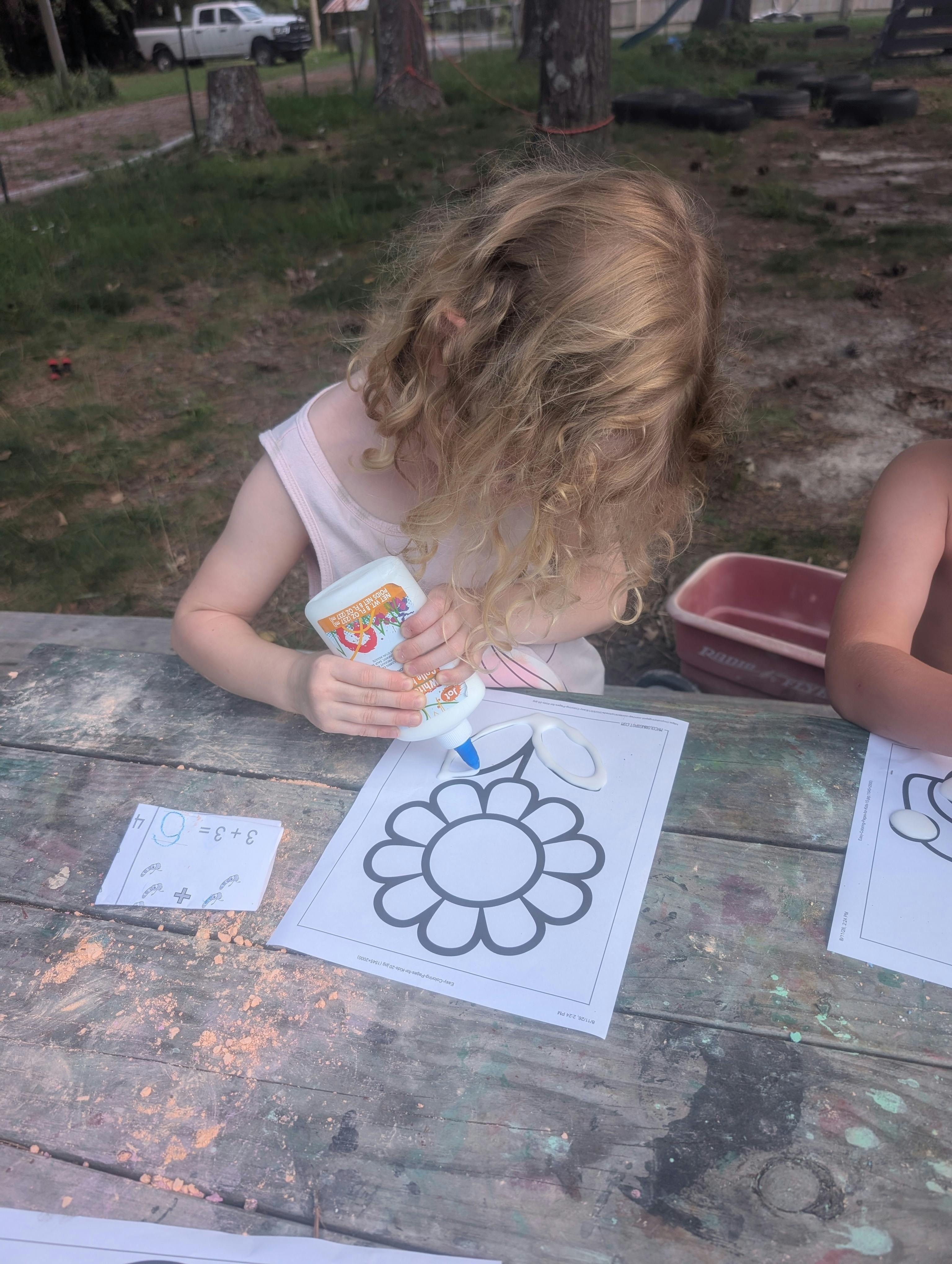 Child applying glue to a flower coloring page at an outdoor wooden table