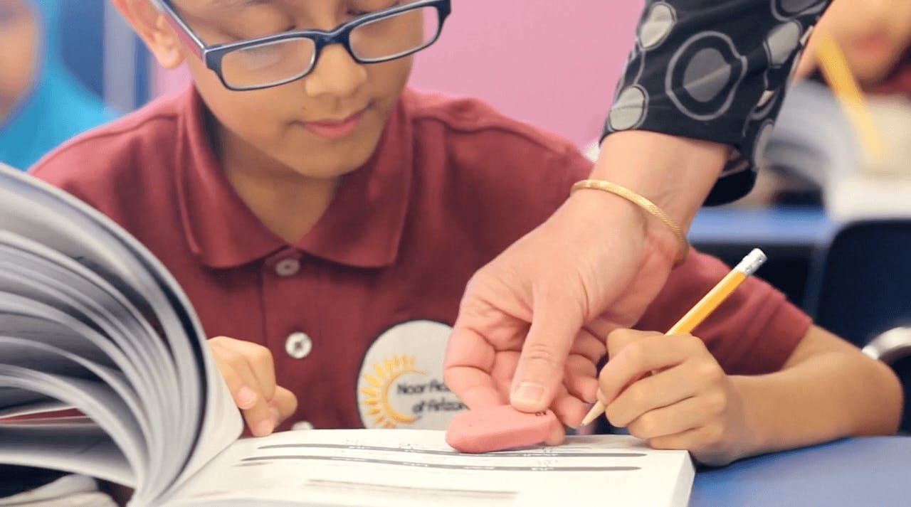 Child in glasses reading a book with an adult hand holding an eraser