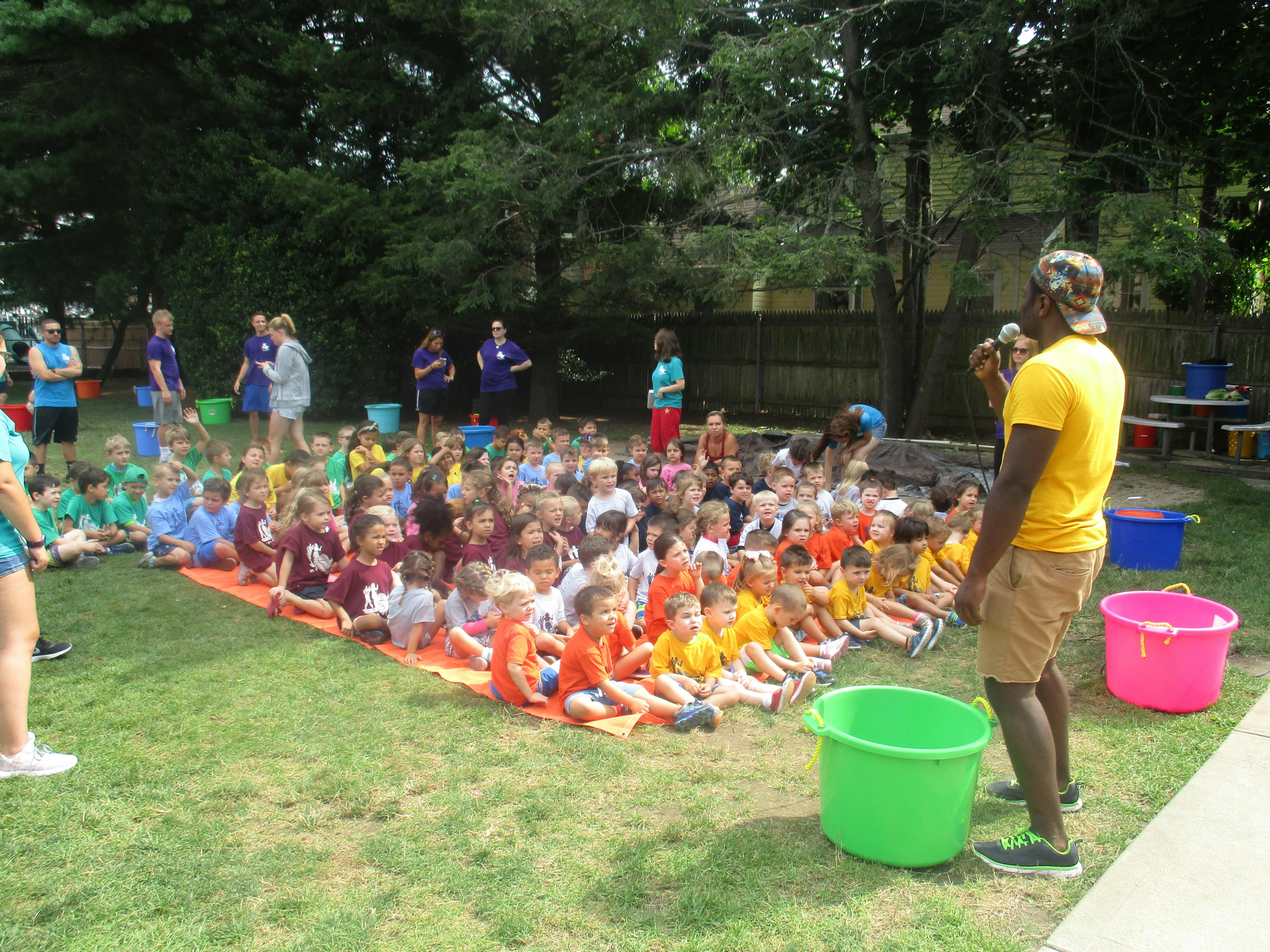 Person speaking to a group of children sitting on a mat outdoors