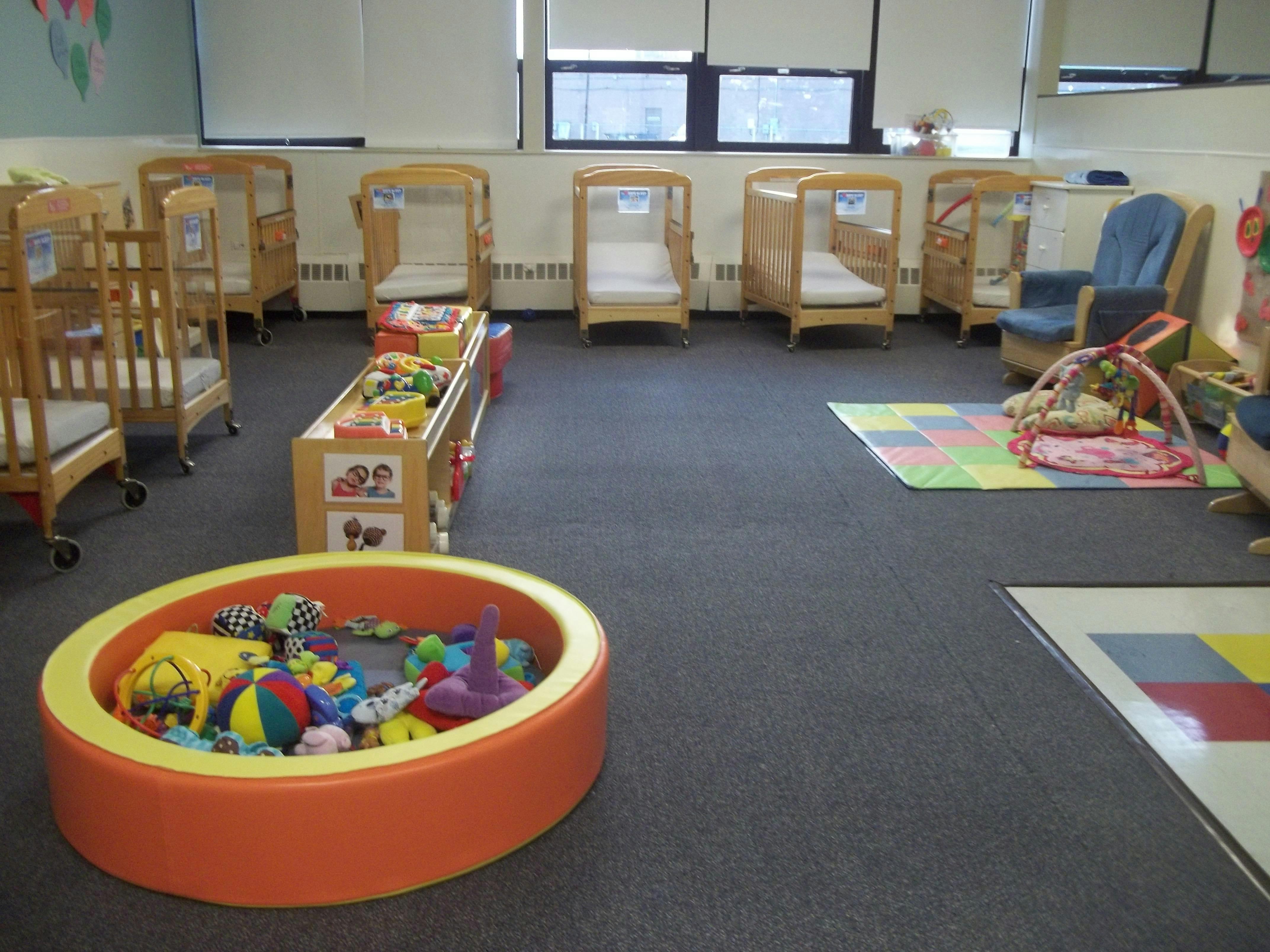 A daycare room with many empty cribs, play mats, and a soft playpen full of toys