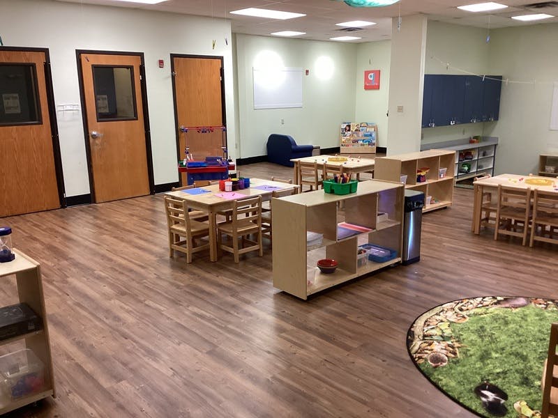 Classroom with several tables and chairs, shelves with toys, and a rug with a nature scene