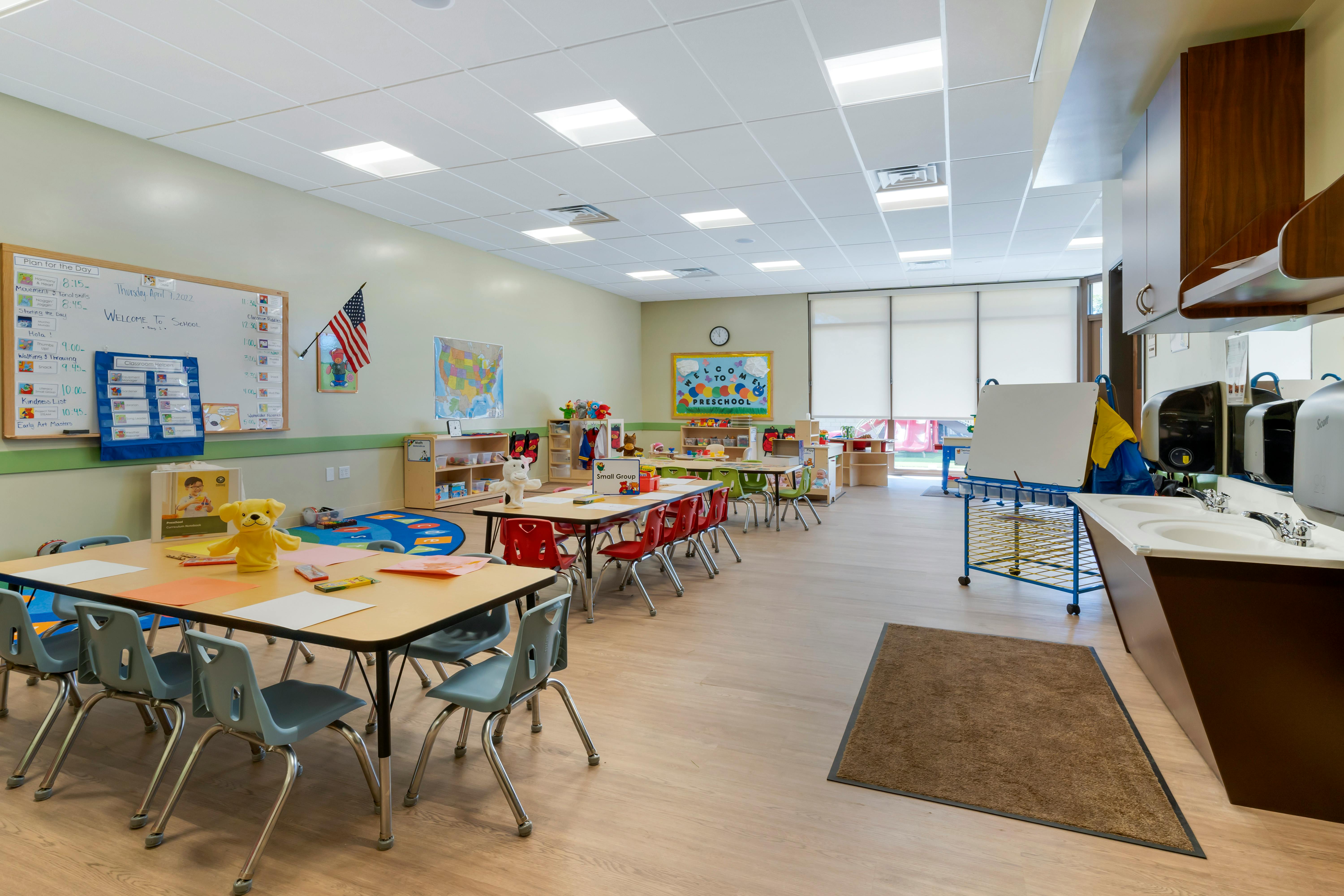 A classroom with small tables and chairs, a sink, and a whiteboard with "WELCOME TO SCHOOL"