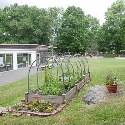 Raised garden bed with plants and metal hoops, building and playground in background