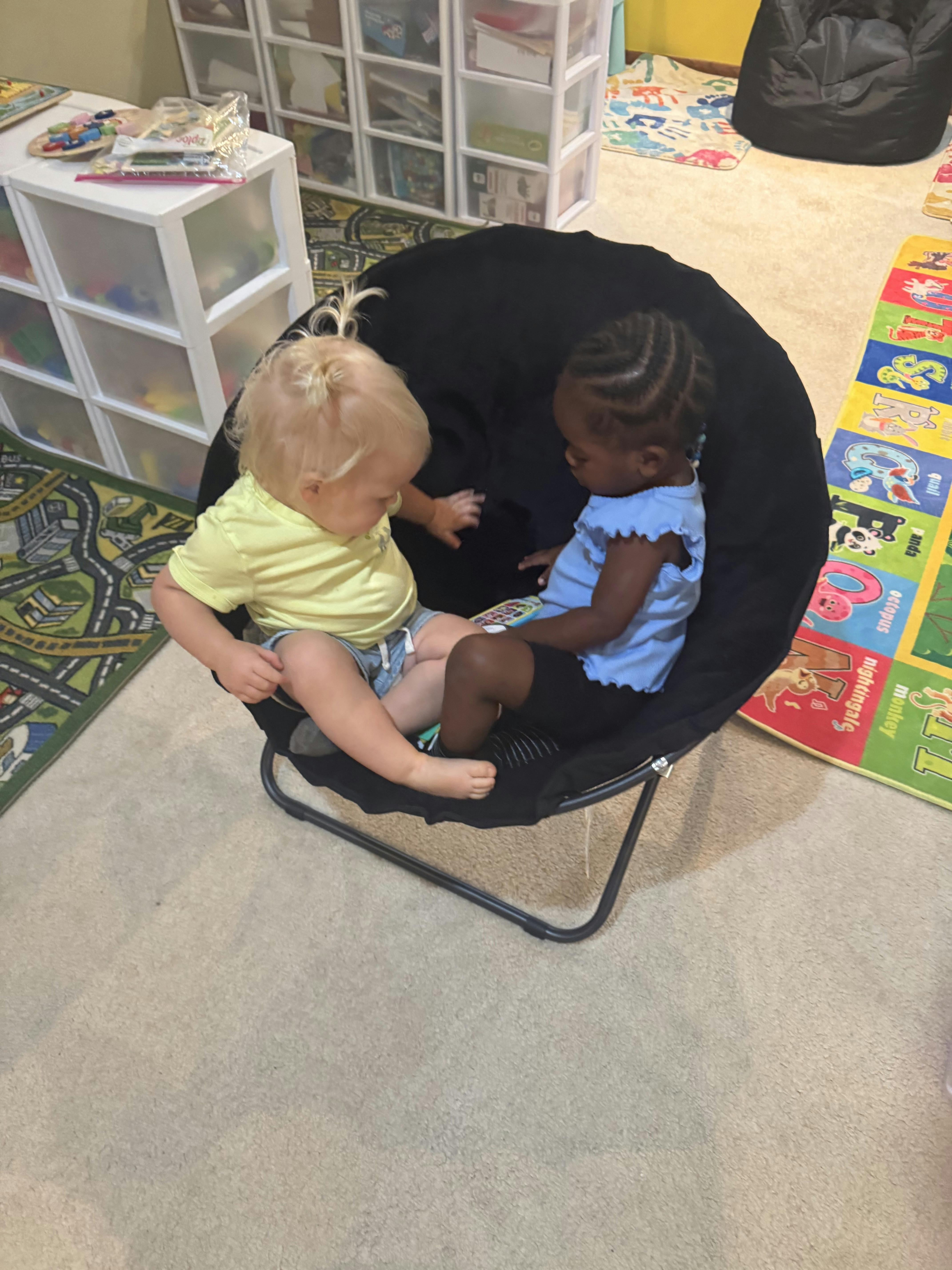 Two children sitting together in a small black saucer chair