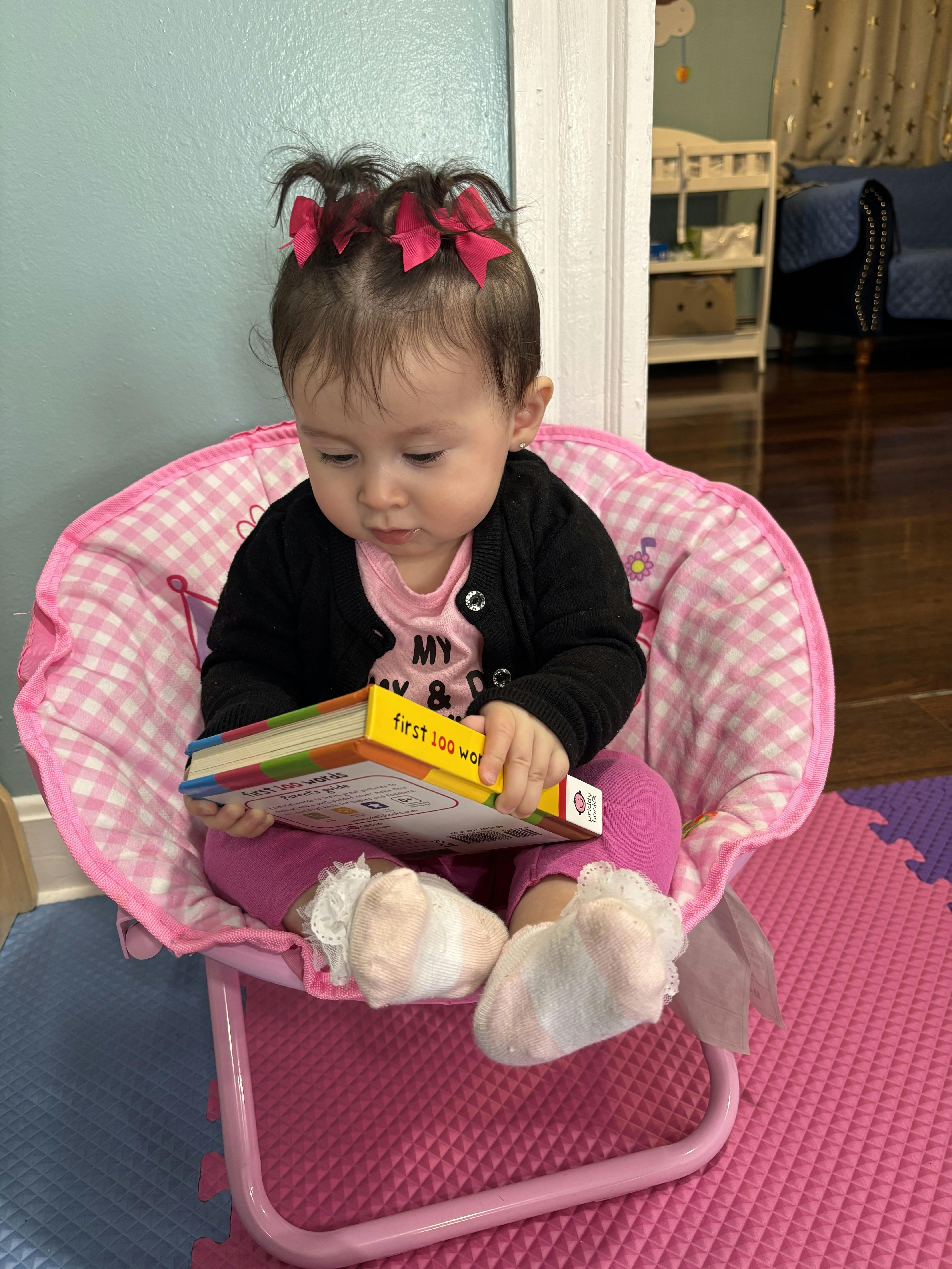 Child sitting in a pink chair holding a book that says "First 100 Words"
