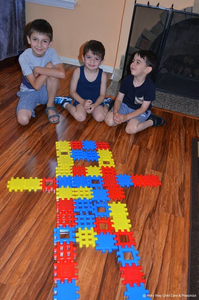 Three children kneeling on a floor with a large toy made of colorful interlocking blocks