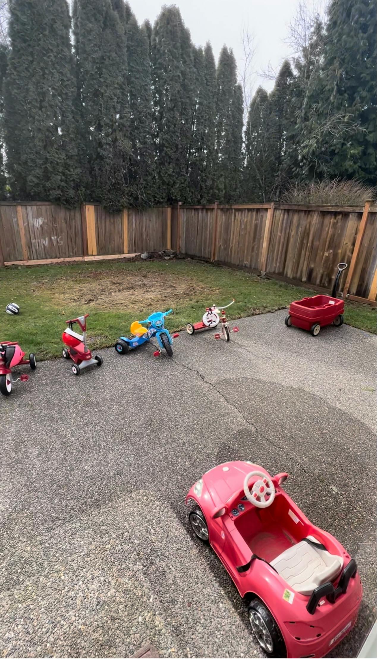 Children's ride-on toys on a paved area next to a wooden fence and trees