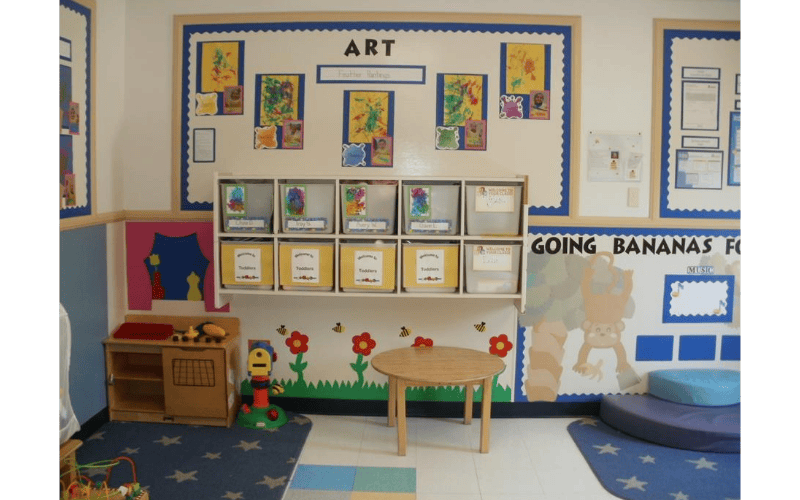 A classroom corner with art displayed on the wall, storage bins, and a small table