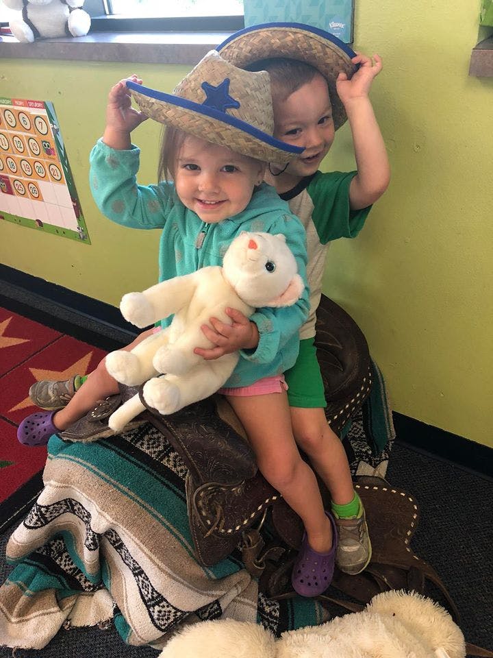 Two children wearing cowboy hats, one holding a stuffed animal, sitting on a saddle