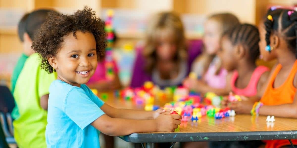 Child smiling at camera while other children play with blocks at a table