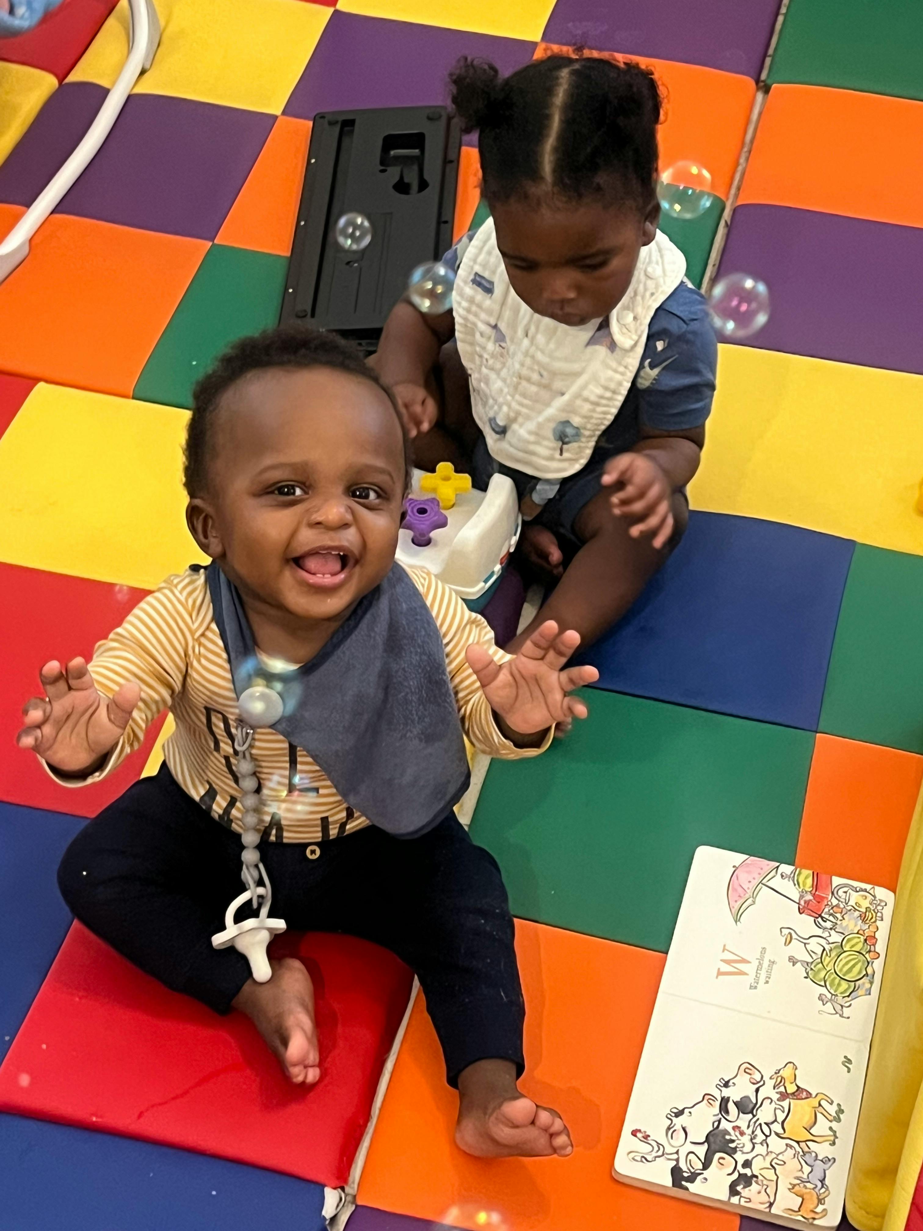 Two children sitting on a colorful mat, one smiling at the camera