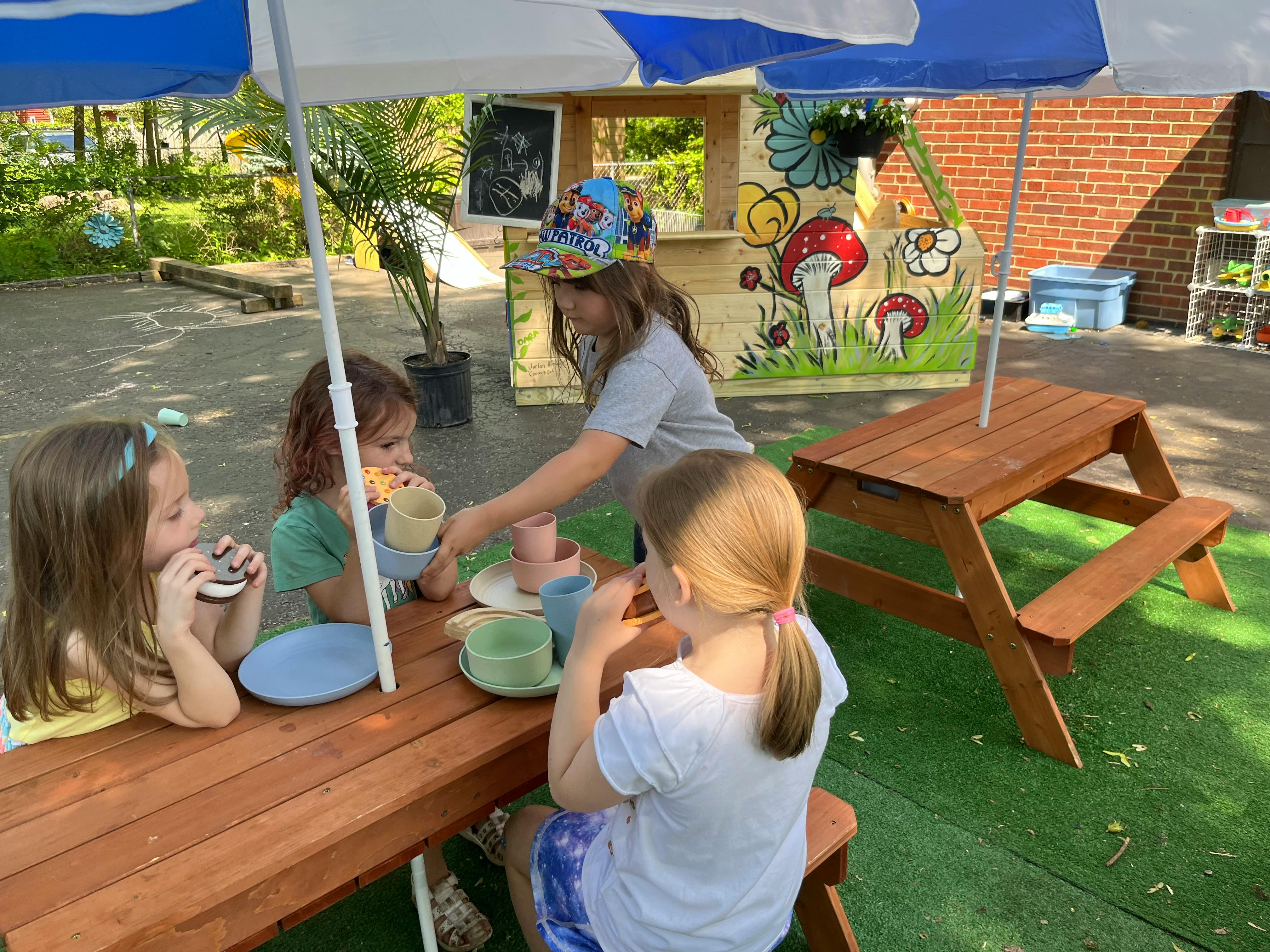 Children playing with toy dishes at a picnic table under an umbrella