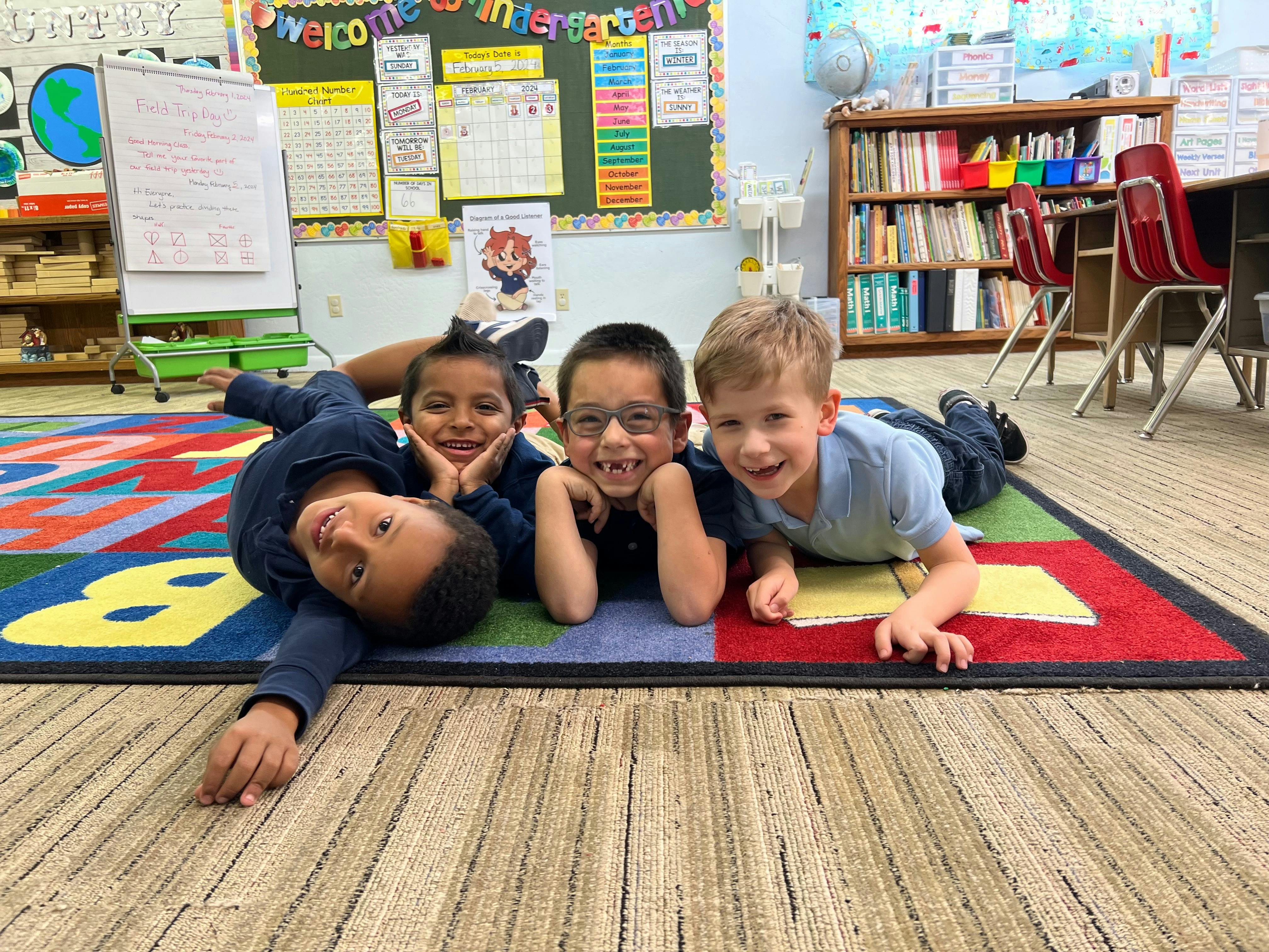 Four children lying on a colorful rug in a classroom