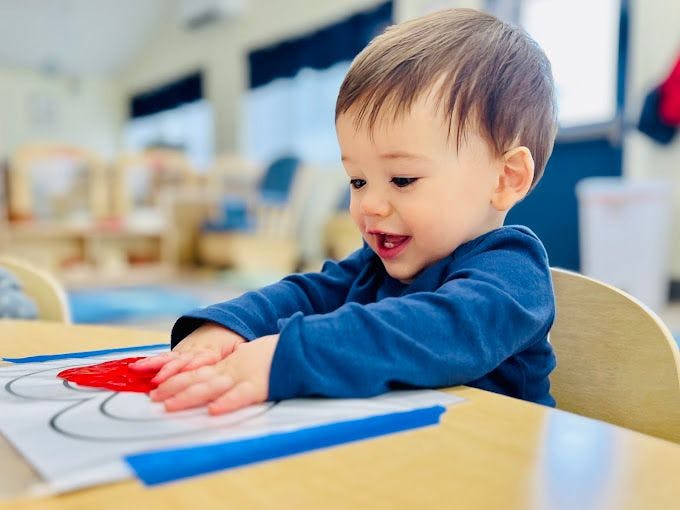 Child pressing hands into red paint on paper at a table in a classroom