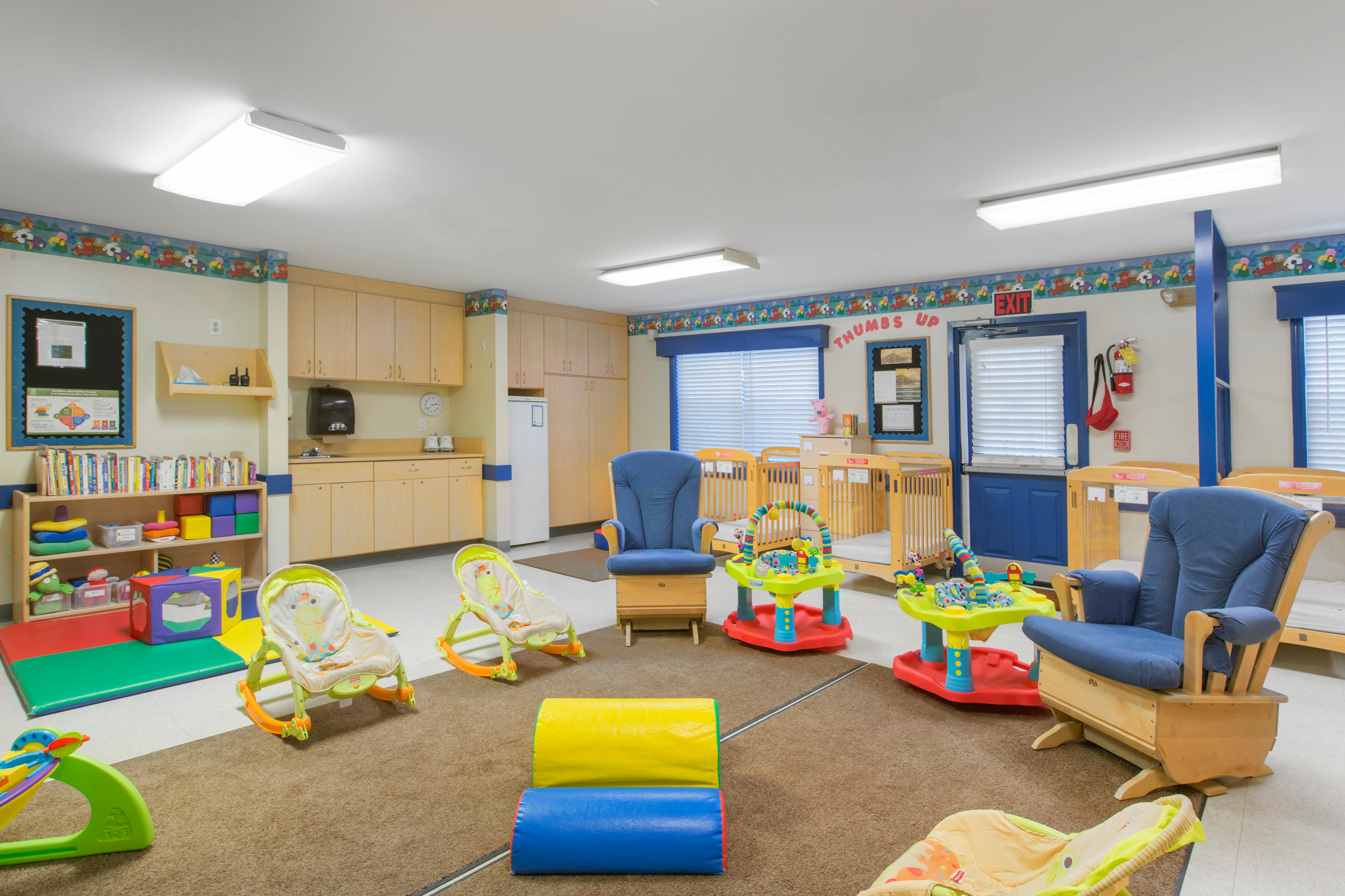 A childcare room with cribs, rocking chairs, baby bouncers, and play mats