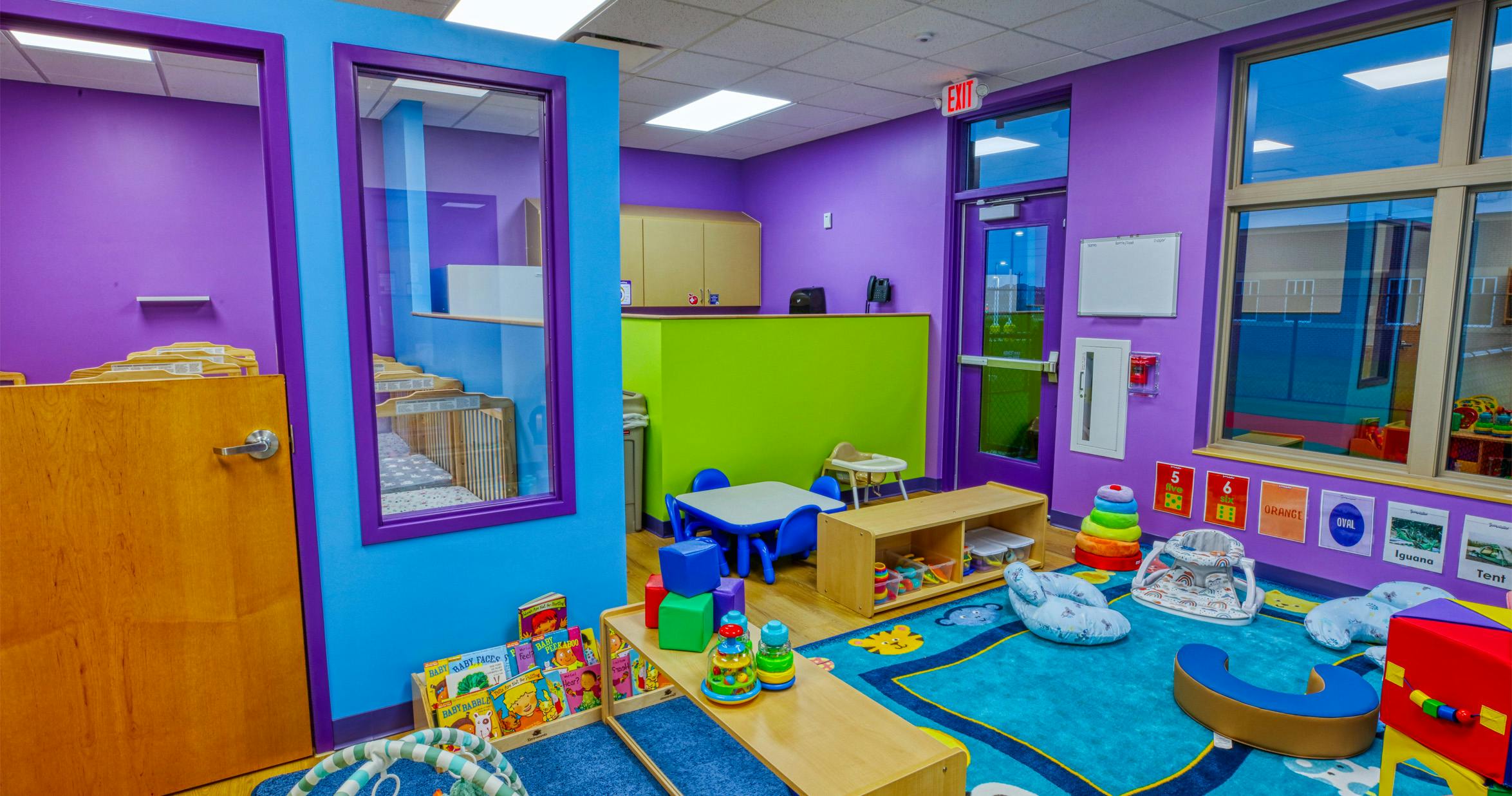 Colorful playroom with cribs visible through a glass wall, and play items on a blue rug