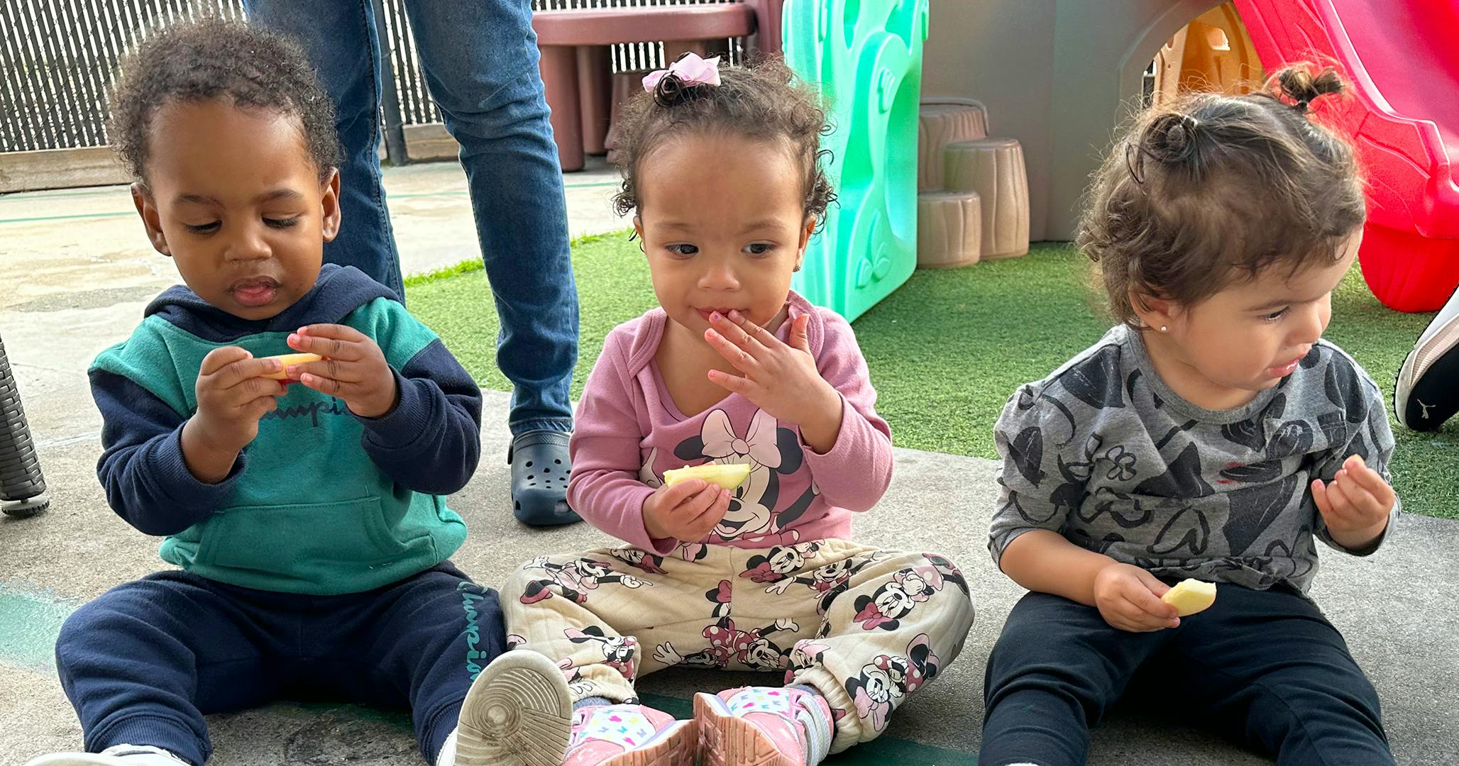 Three children sitting on the ground eating apple slices on a playground