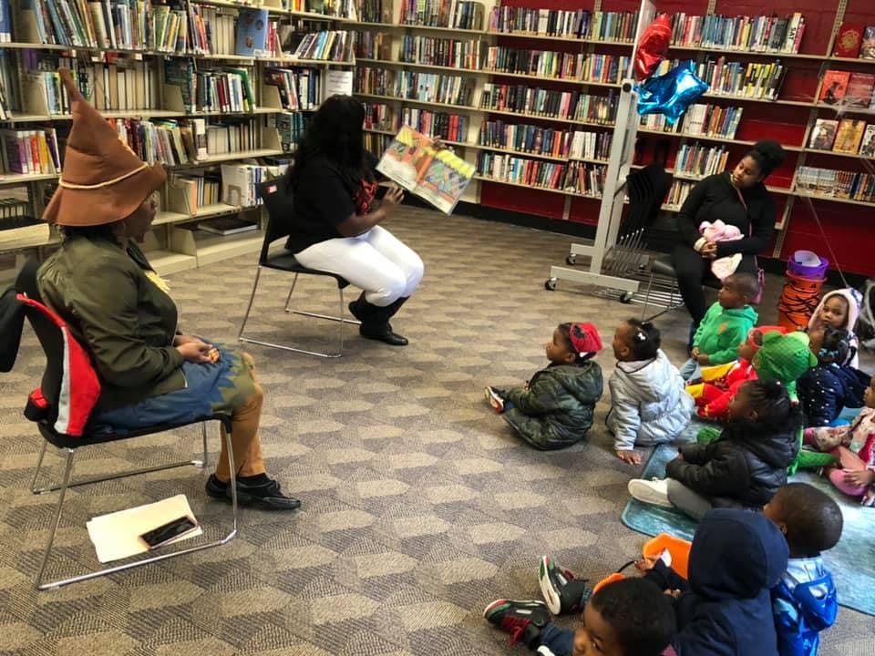 Person in a hat reading a book to children in a library