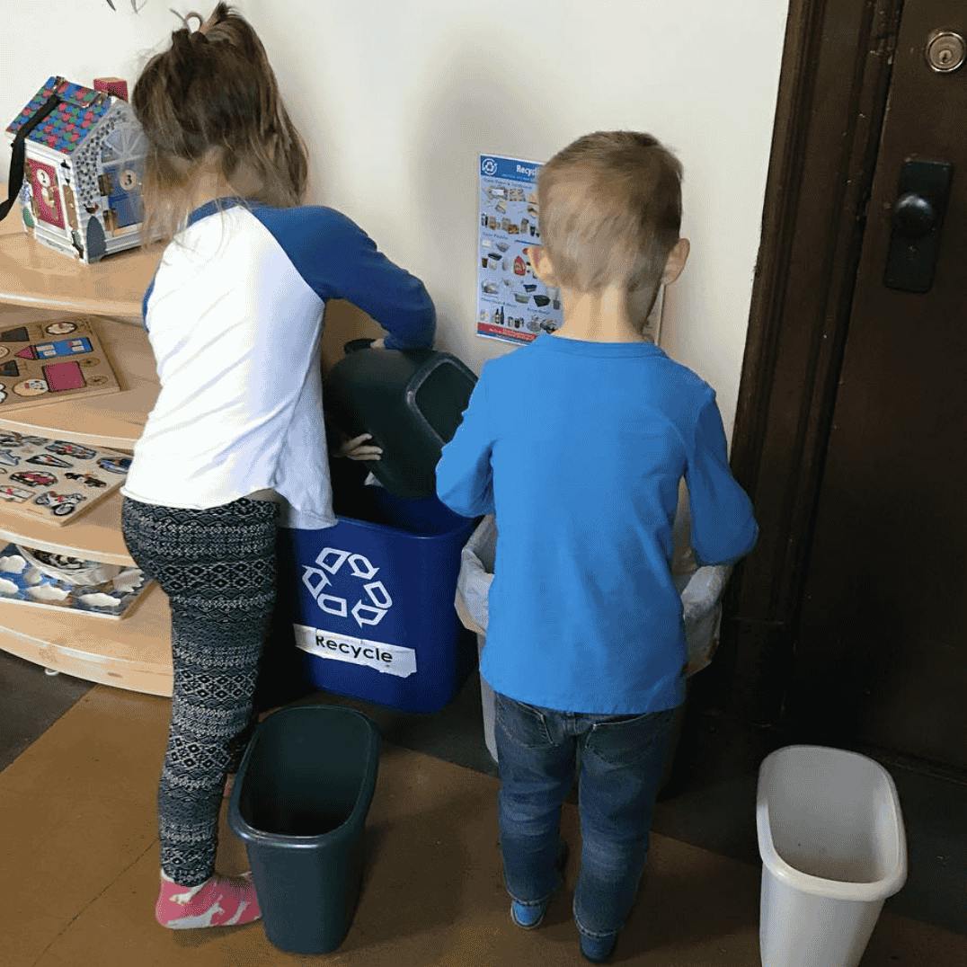 Child dumping a small bin into a recycle bin while another child watches