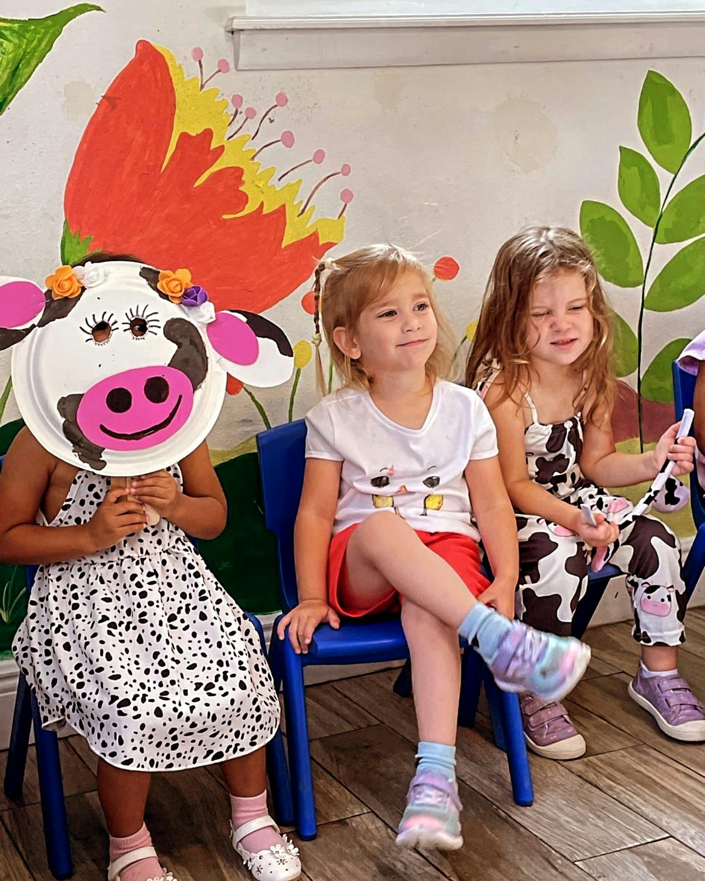 Three children sitting on chairs, one holding a cow mask to their face