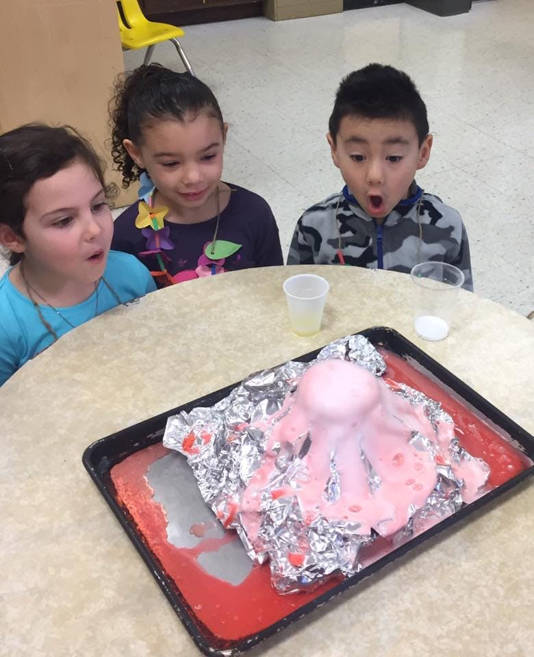 Three children watching a science experiment with a pink volcano erupting