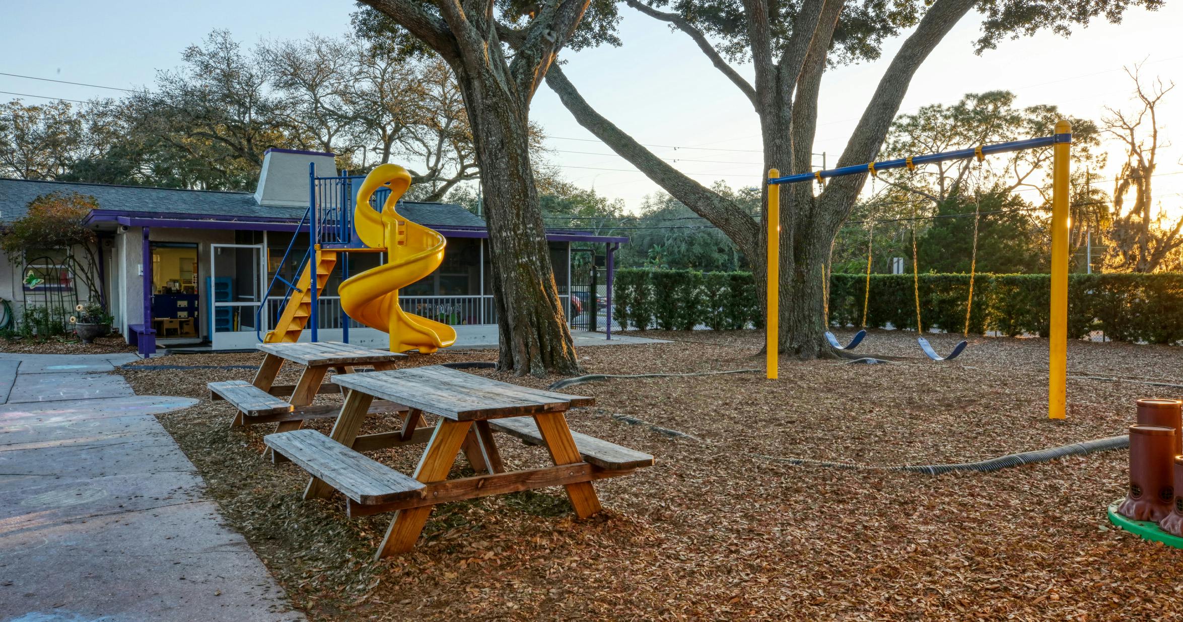 Empty playground with a yellow slide, swings, and picnic tables next to a building