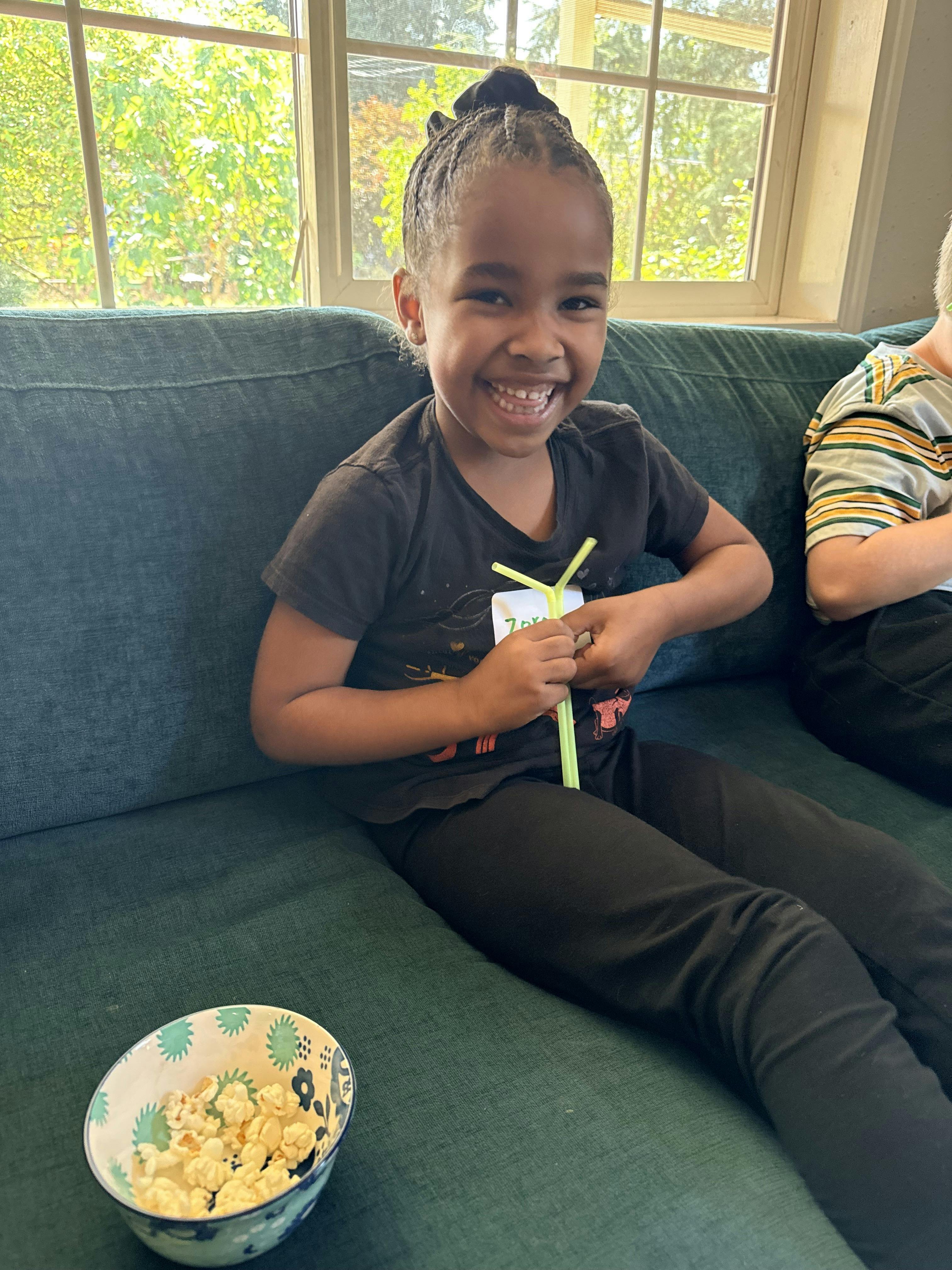 Child sitting on a sofa holding a straw next to a bowl of popcorn